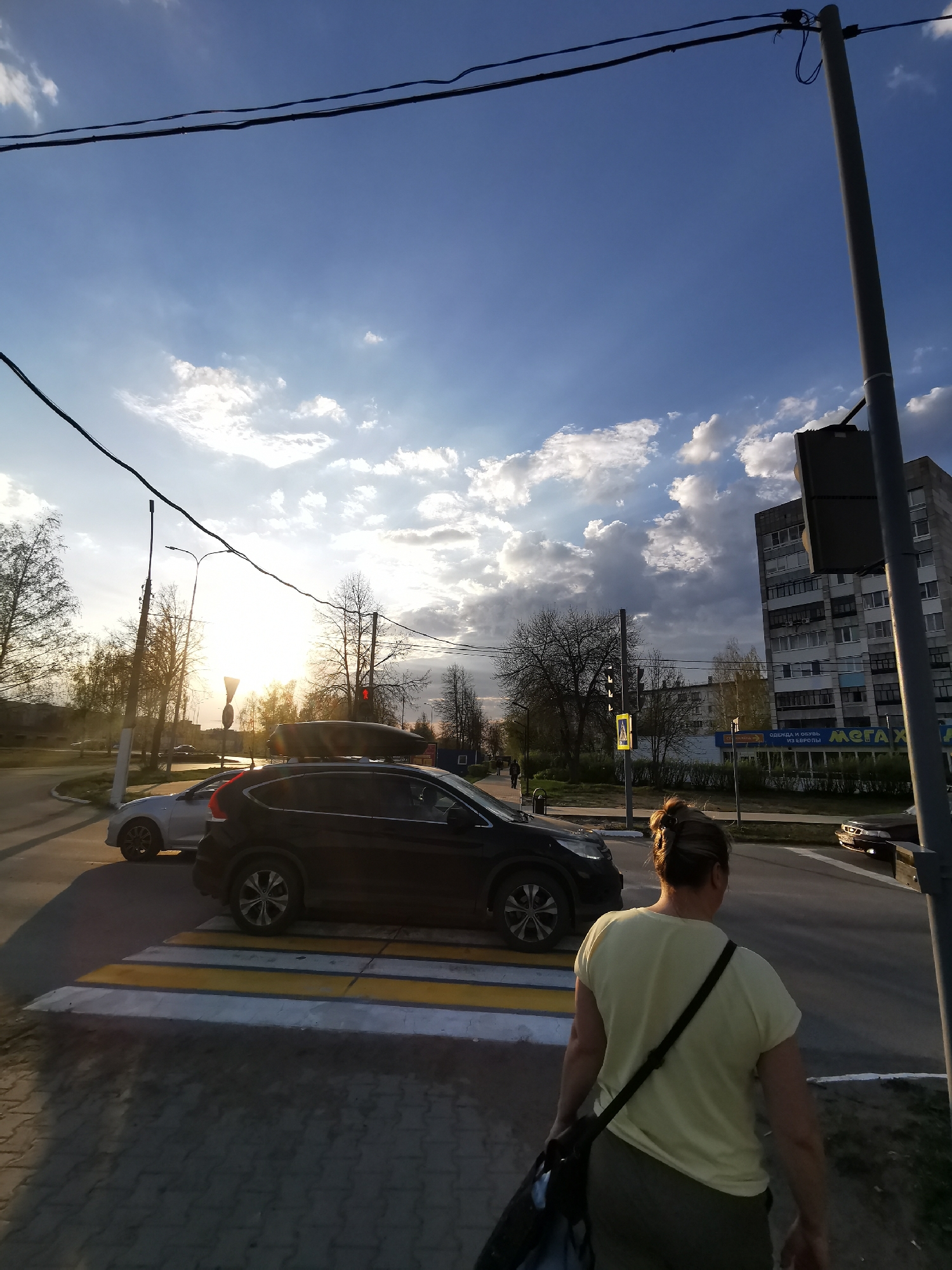 The photograph depicts a street scene in the city of Unknown, Unknown, taken during daylight. A woman in a light yellow t-shirt and dark trousers is walking away from the camera, across a zebra crossing. A black car is in the middle of the crossing, and a silver car is further back. A multi-story building is visible in the background, adjacent to a traffic light. The sky is partly cloudy, and the sun is setting, creating a bright glow in the distance. Power lines stretch across the upper portion of the frame. A streetlight and a traffic light pole are present on the right side of the image.