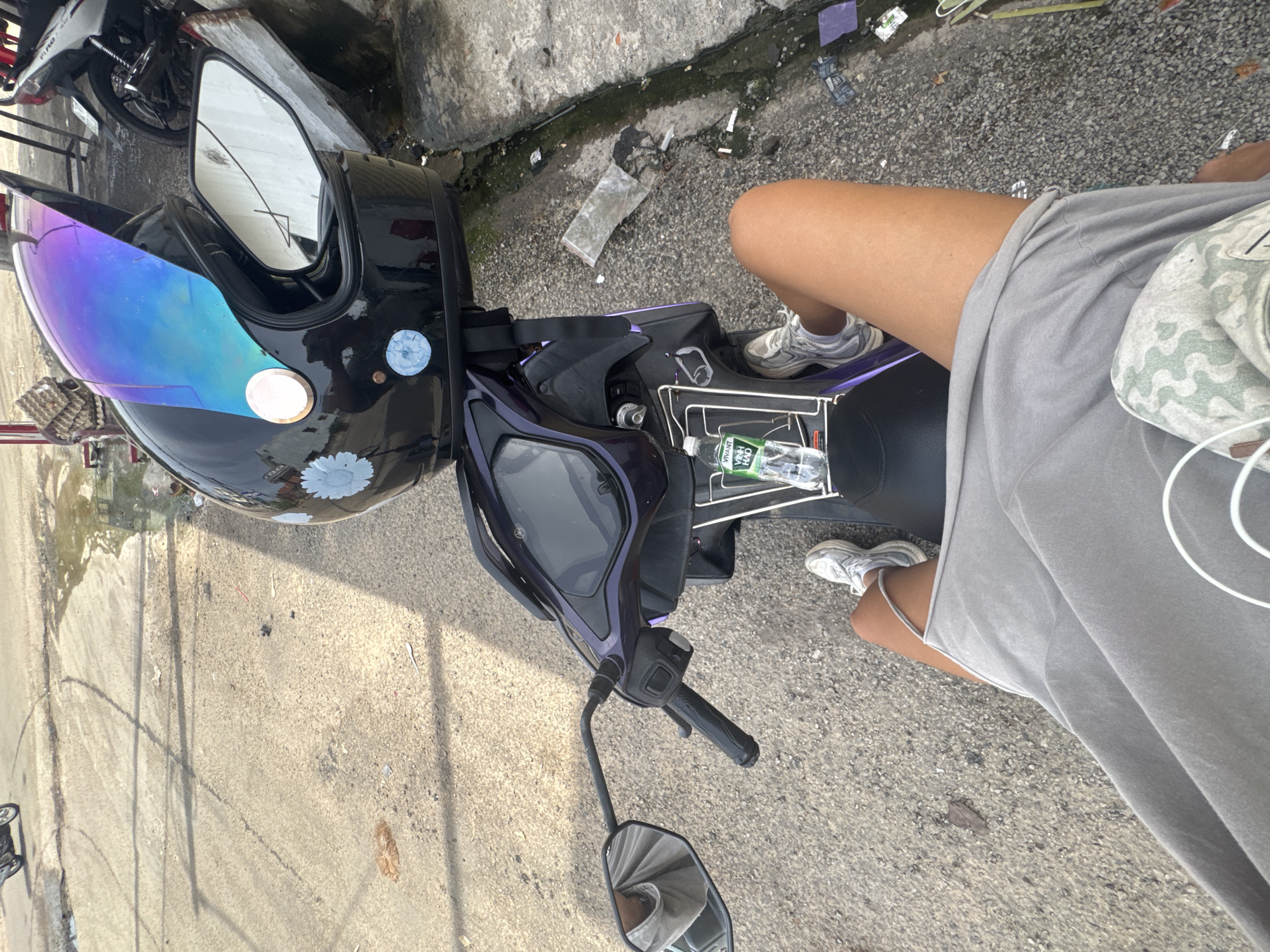 The image shows a first-person perspective of someone sitting on a dark purple motorcycle. The rider is wearing grey shorts and white sneakers, with a grey t-shirt visible. A black motorcycle helmet with a iridescent visor is on the seat in front of the rider, adorned with two light blue flower stickers. A partially visible white bag with a green zigzag pattern is also on the seat. A plastic bottle of "VIVANT" brand water with Vietnamese text is clipped to the side of the motorcycle.
The motorcycle is parked on a paved surface next to a concrete sidewalk and a drainage ditch filled with debris and some water. In the background, a portion of another motorcycle, a white one, is visible. The ground appears to be a mix of asphalt and gravel, with some litter scattered around. The scene suggests an outdoor setting, likely on a street or roadside in a populated area. The lighting indicates it is daytime, with shadows cast on the ground. There are no overt indications of weather conditions beyond the presence of some water in the ditch. The overall impression is a casual stop or pause during a motorcycle journey in a somewhat unkempt urban or semi-urban environment.