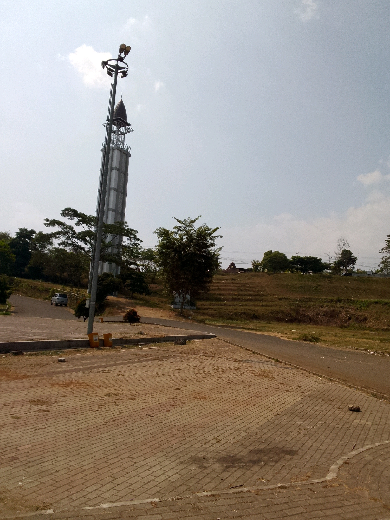 The image depicts an outdoor scene with a tall, slender tower as the central focus, situated in an open area with paved surfaces and grassy slopes. The tower, which appears to be a minaret, has a metallic structure with a pointed finial and is illuminated by a cluster of lights at its apex. To the left of the tower, a gray car is parked on a paved road that curves away into the background. The foreground features a textured brick-paved area, with some scattered debris. Behind the paved area and the tower, a grassy hillside is visible, with trees dotting the landscape. Further in the background, a road winds through the terrain, and a small, rustic building with a triangular roof is perched on a higher elevation. The sky is partly cloudy, indicating daytime. The overall environment suggests a public park or a complex with religious and recreational facilities. This scene is located in Pasirhalang, Indonesia, as indicated by the presence of a minaret, a common architectural feature in the region.