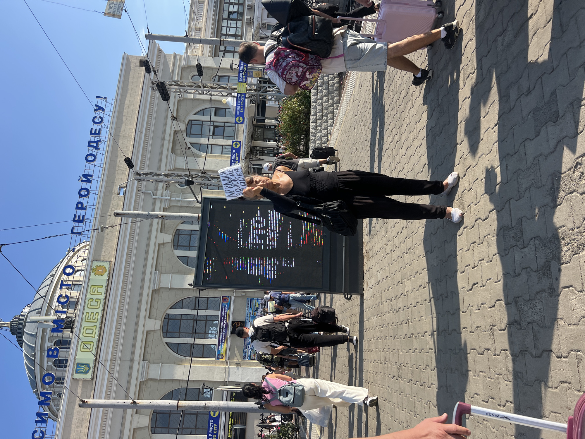 An outdoor, eye-level, medium shot shows a woman holding a sign at the Odesa railway station, Ukraine. The woman, in the foreground, is wearing a black tank top and black pants. She holds a sign that says "АРКАДІЯ ЗОЛОТА БІЛКА" in Ukrainian. A large digital display board is to her left, showing colorful text. Behind her, people with luggage walk on a paved walkway in front of the station building. The station has large arched windows and a dome. The sky is clear and blue.