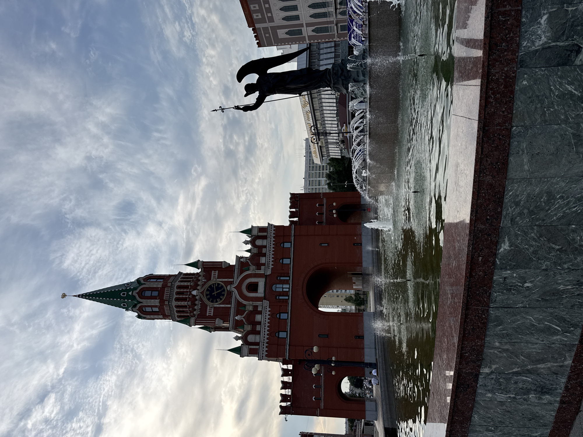 The image shows the Monument of the Archangel Michael in Yoshkar-Ola, Russia. The statue is located in a plaza with a fountain in front of it. In the background, there is a red brick tower with a clock and a green spire, which is part of the National Art Gallery. The sky is cloudy with some patches of blue. There are several people sitting on the edge of the fountain, and some are walking in the background. The fountain is active with water jets. The overall scene depicts a public space with historical and artistic elements.