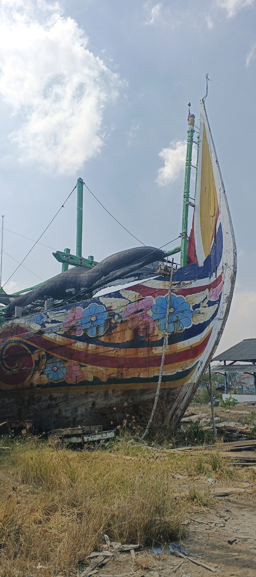The image shows the front of a wooden boat, possibly a fishing vessel, pulled ashore in the city of Blimbing, Indonesia. The boat is heavily decorated with colorful, faded paintings of flowers and stripes along its hull. It appears weathered and somewhat dilapidated, with some parts of the paint peeling. A black net covers a portion of the boat. Two green masts rise from the deck, with some rigging attached. The boat rests on dry, grassy ground with scattered debris. In the background, a simple structure with a dark roof is visible, along with more trees and buildings in the distance. The sky above is blue with scattered white clouds. The scene suggests the boat is either undergoing repairs or has been decommissioned. There are no people visible in the image.