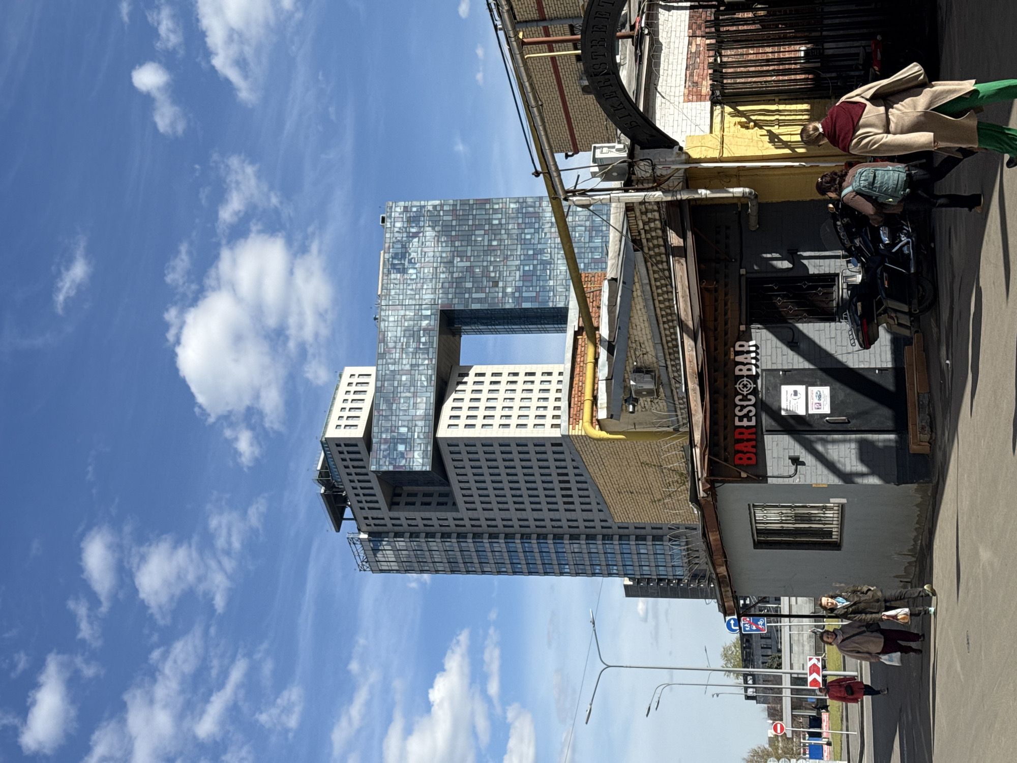 The image depicts a vibrant urban scene in Moscow, Russia, captured on a sunny day with a clear blue sky dotted with white clouds. The photograph is oriented vertically, showing the sky at the top and the street level at the bottom.
In the mid-ground and dominating the left side of the frame is a distinctive, modern skyscraper. This tall building features a striking design with a large, rectangular void or cutout in its upper section. Its facade is a mosaic of varying blue, grey, and white reflective glass and opaque panels, creating a dynamic, patterned appearance. Below this, simpler, light-colored sections with multiple small windows are visible.
To the right, contrasting with the modern high-rise, are older, lower-rise brick buildings. One building, with painted grey walls and exposed yellow and reddish-brown brickwork above, features a prominent red sign that reads "BAR ESCO BAR" with a target symbol. Above this, a black, curved sign is partially visible, displaying the text "BIKER'S STREET". Various utility pipes and electrical conduits run along these structures.
Several people are present in the scene. Near the "BAR ESCO BAR" building, one person in a beige coat and green trousers stands, while another person with a light blue backpack appears to be interacting with a parked black motorcycle. Further down the street on the right, two more individuals are walking on the pavement, one in a brown jacket and another in a red jacket.
The street level features typical urban infrastructure, including traffic signs (a blue circular sign, a red triangular sign with a curved arrow, and a red circular 'no entry' sign) and streetlights with multiple arms. The bright daylight casts clear shadows, indicating midday to late afternoon. The combination of modern architecture and older, more utilitarian buildings provides a visual contrast characteristic of many evolving urban centers.