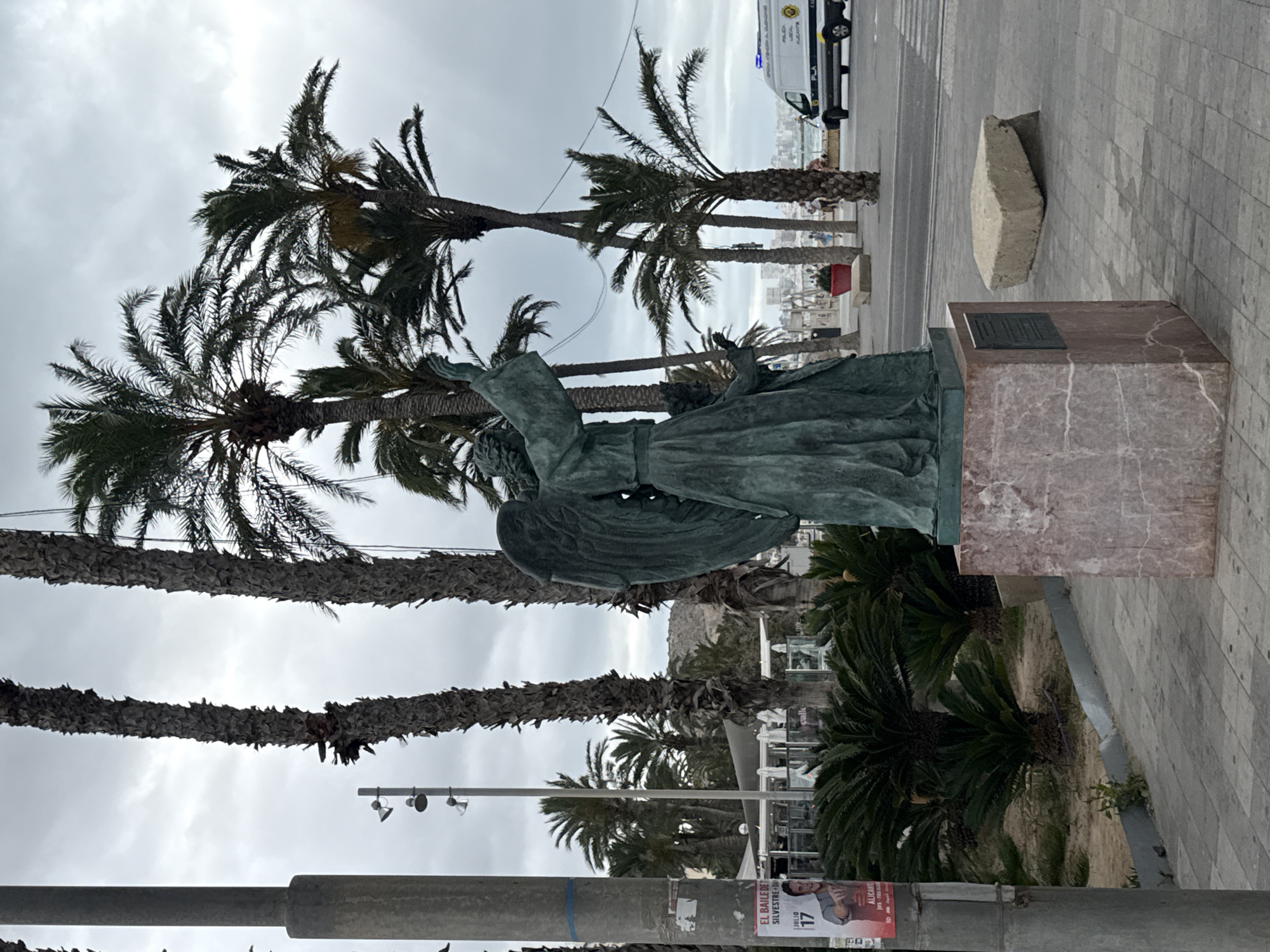 The image depicts a statue of an angelic figure in an outdoor, coastal setting under an overcast sky. The statue, made of weathered bronze or copper, features large wings, flowing drapery, and outstretched arms, with its gaze directed upwards and slightly to the left. It stands on a reddish-brown marble pedestal that includes a rectangular dark plaque on its upper surface. To the right of the pedestal, a large, irregularly shaped light-colored stone block rests on the patterned grey pavement.
Numerous tall palm trees with textured trunks and feathery fronds are prominent throughout the scene, indicating a warm climate. In the background, beyond the palm trees, a body of water (likely the sea) and distant buildings suggest a city coastline. A white police van, marked with "POLICIA LOCAL ALICANTE" and "PLA," is visible on a paved area, with a few indistinct people nearby.
In the foreground, a utility pole or lamppost runs vertically along the left. Attached to it is a poster with partially visible text, including "EL BAILERE DE," "SILVESTRE D...", "JULIO 17," and "ALICANTE." The scene is set during the daytime, characterized by a grey, cloudy sky.