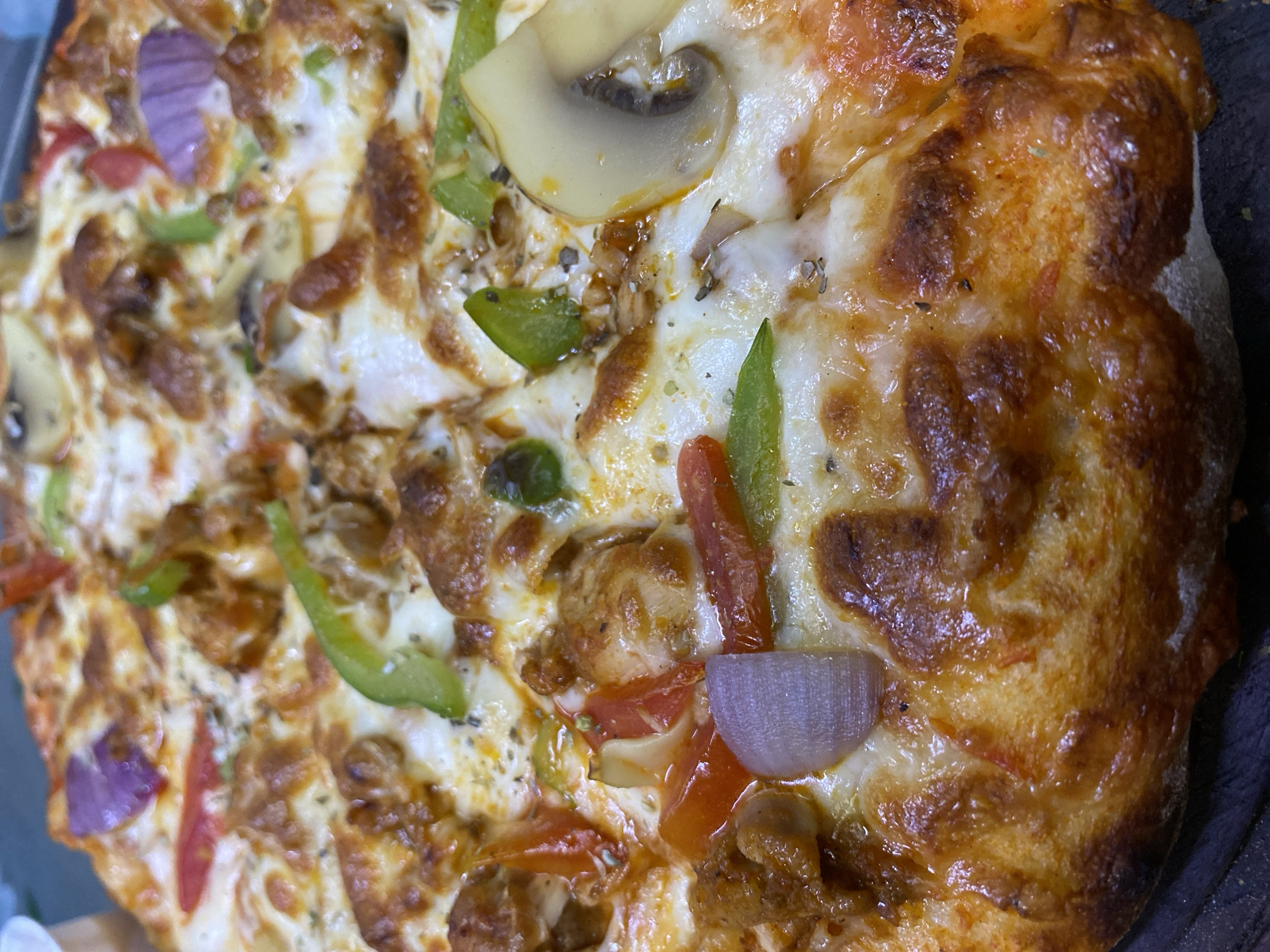A close-up shot of a pizza with a variety of toppings, including mushrooms, green peppers, red onions, tomatoes, and what appears to be chicken or meat chunks. The cheese is melted and bubbly, and the crust is golden brown. The pizza is cut and appears to be served on a dark-colored surface. No specific location is identifiable from the image to confirm its location as Dhaka, Bangladesh. No people are visible in the shot.