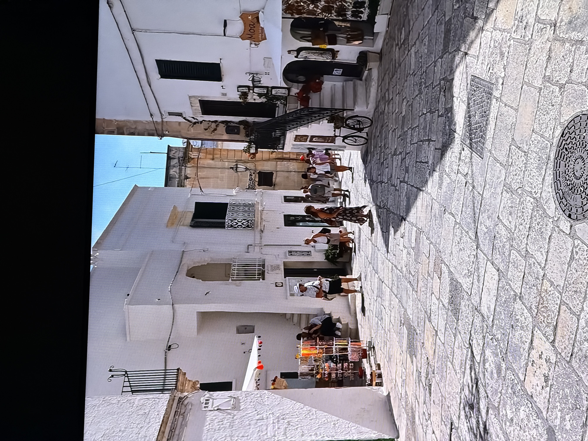 This is a photograph of a narrow street lined with whitewashed buildings. The street is paved with large, irregular stone slabs. Several people are walking and standing in the street. On the left, a rack of colorful jewelry and accessories is displayed outside a shop. Further down the street, people are seated on steps. On the right, a white building has a shop with displays of goods, and a bicycle is parked next to an external staircase. The architecture is typical of a Mediterranean town, with white walls, arched doorways, and balconies with decorative railings. The sky is bright blue, indicating a sunny day. The scene suggests a bustling marketplace or a tourist area. The photograph is framed by a black border, implying it might be a picture displayed on a screen.