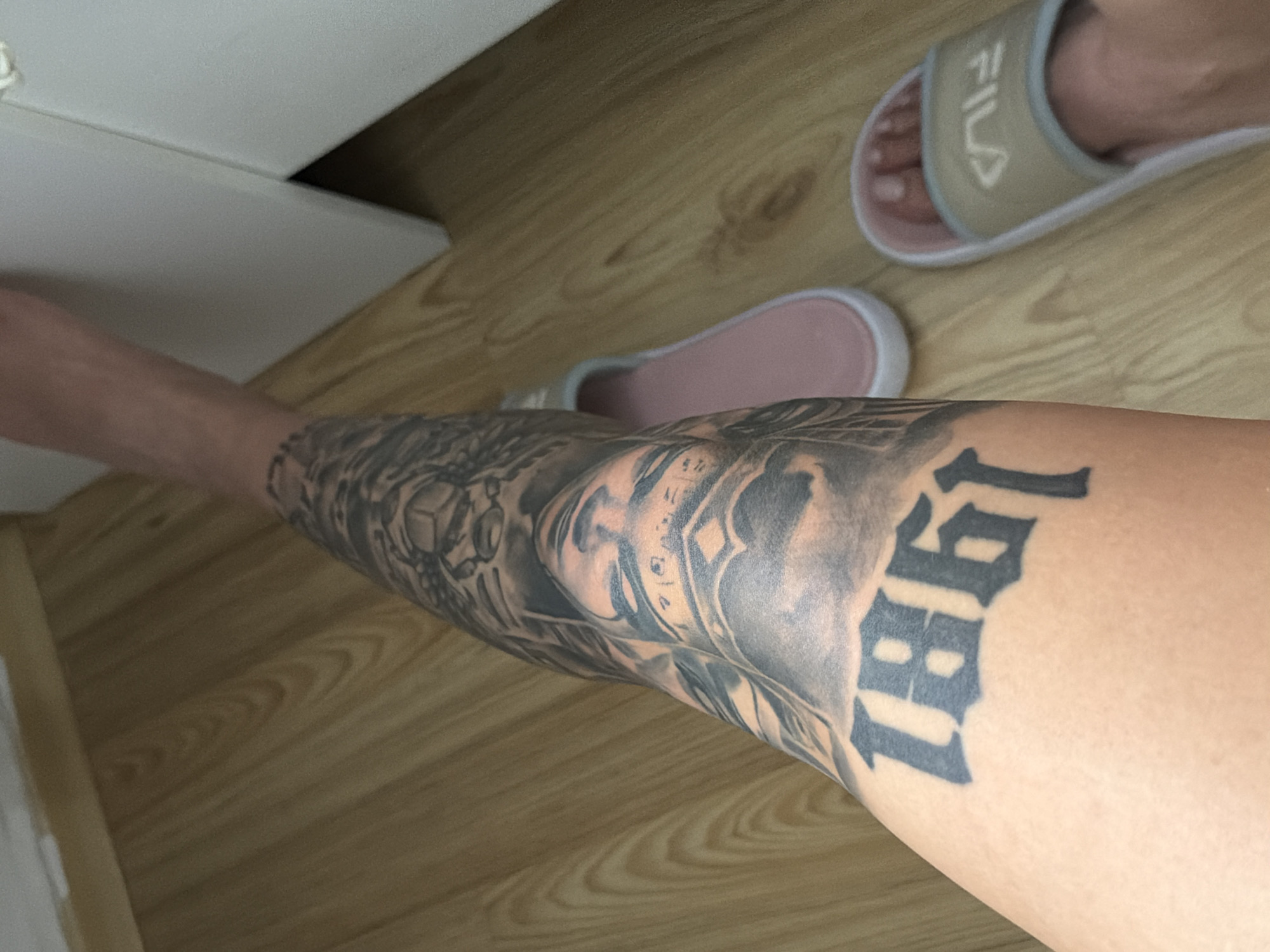 A person's leg with a large black tattoo of the numbers "1861" is visible against a wooden floor. Two pink and white slide sandals are on the floor near the leg, one with a Fila logo. The scene is indoors. There is no information in the image to determine the location in Plovdiv, Bulgaria.
