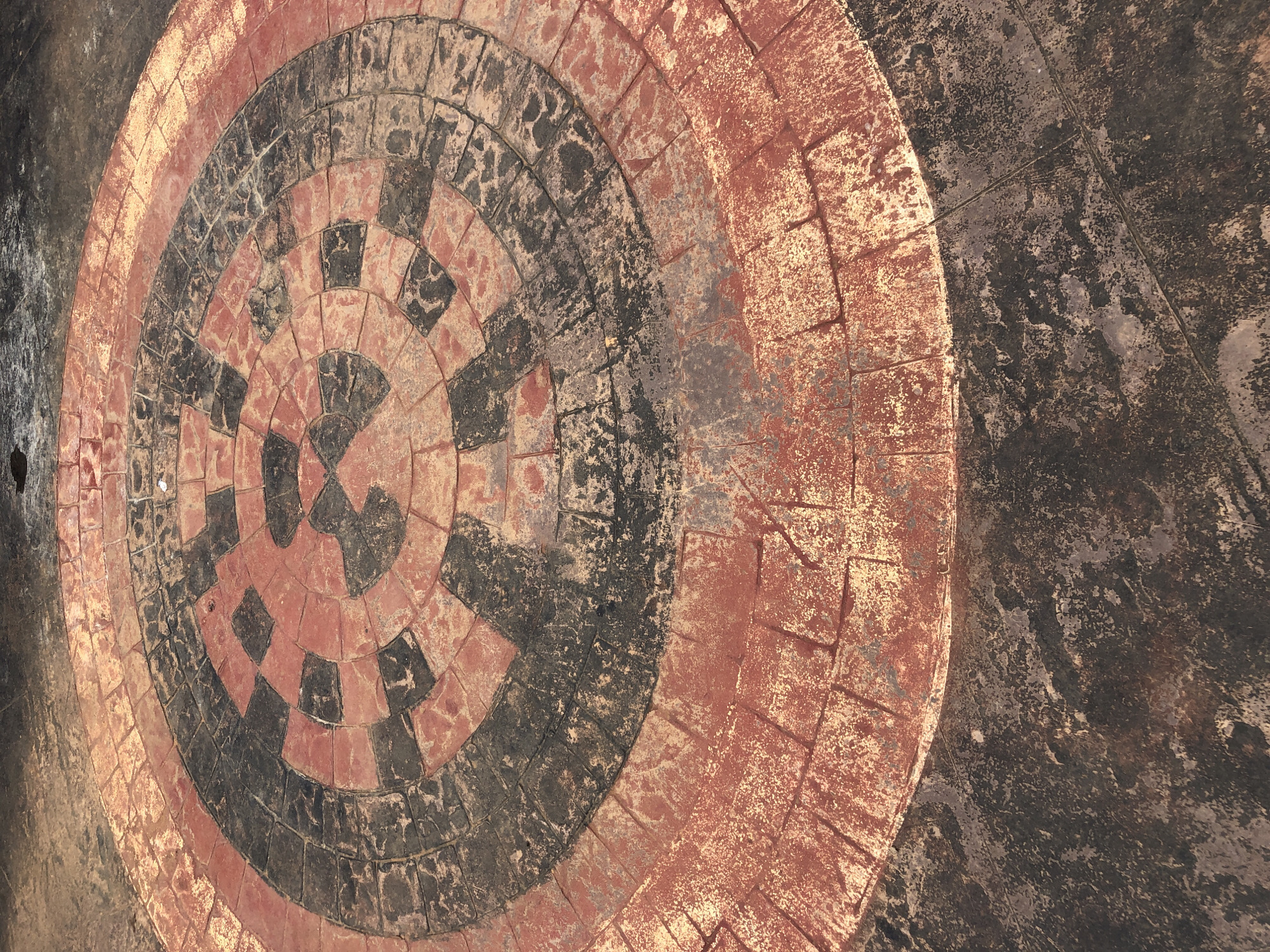 The image displays an overhead view of a circular, patterned ground surface. The central design is composed of concentric rings made of small, rectangular blocks, resembling paving stones or tiles. These blocks alternate in color between a weathered reddish-orange and a dark grey or black, forming a segmented, checkerboard-like pattern that spirals outwards. The outermost visible ring of the circular pattern is primarily reddish-orange. This patterned area is set into a larger, darker, and rough-textured surface, likely concrete or asphalt, which fills the surrounding space of the frame.
The entire surface appears aged and worn, with the colors showing signs of fading, scuffing, and accumulation of dirt or grime, suggesting long-term exposure to outdoor conditions. Both the reddish-orange and dark blocks exhibit variations in shade and texture due to wear and environmental factors. There is no activity or event taking place, as the image focuses solely on the static ground pattern. No people or specific objects are visible. The lighting suggests an outdoor daytime setting, but the exact time of day or specific weather conditions are not clear. There are no visible location cues, explicit emotions, or any discernible text in the media file.