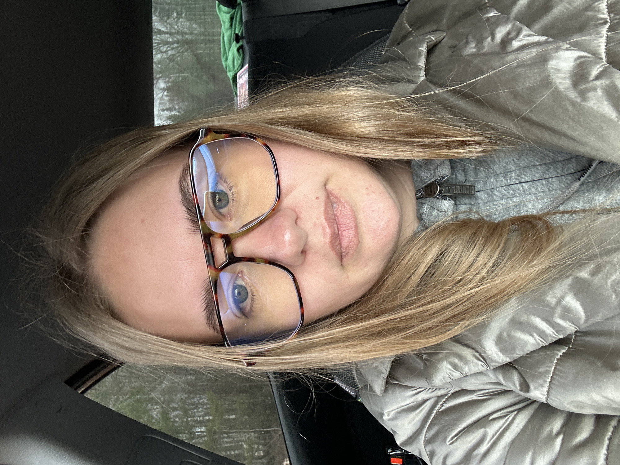A blonde-haired person wearing glasses is positioned in what appears to be the driver's seat of a vehicle. The person is wearing a gray jacket. The background shows trees through the car window. The location is indiscernible.