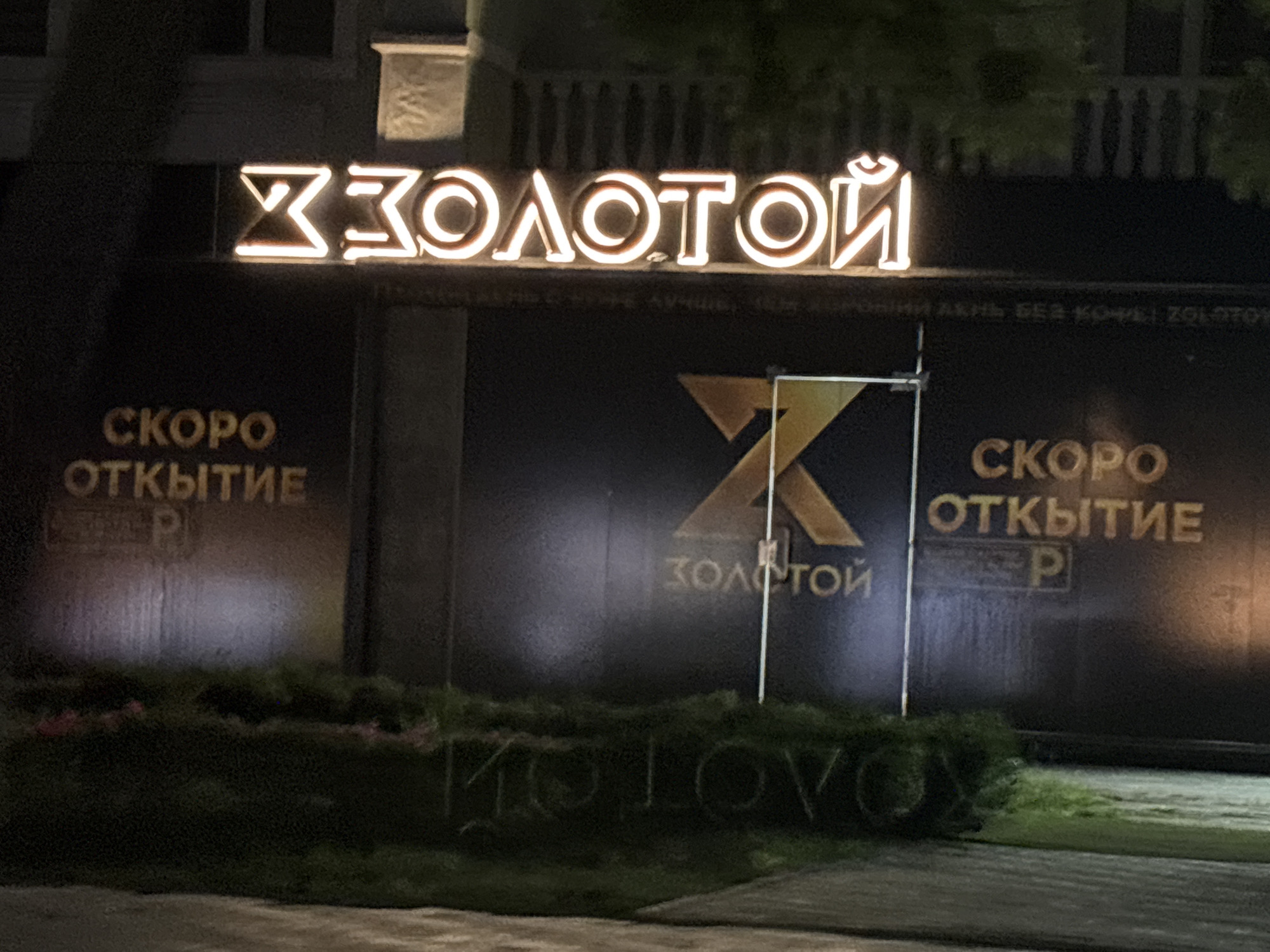 The image shows a nighttime view of a building facade in Grozny, Russia, featuring illuminated signage. The primary sign displays the word "ЗОЛОТОЙ" (ZOLOTOY) in large, neon-like letters, with a stylized "X" logo above it. Below this, on either side, are signs that read "СКОРО ОТКРЫТИЕ" (SKORO Otkrytie), meaning "Opening Soon," with a "P" indicating parking. The building itself appears to be a commercial establishment, possibly a store or entertainment venue. The lighting creates a dramatic effect, highlighting the signs against the dark background. The foreground shows a patch of green bushes and a sidewalk, suggesting an urban setting. The overall impression is one of a newly opened or soon-to-open business.