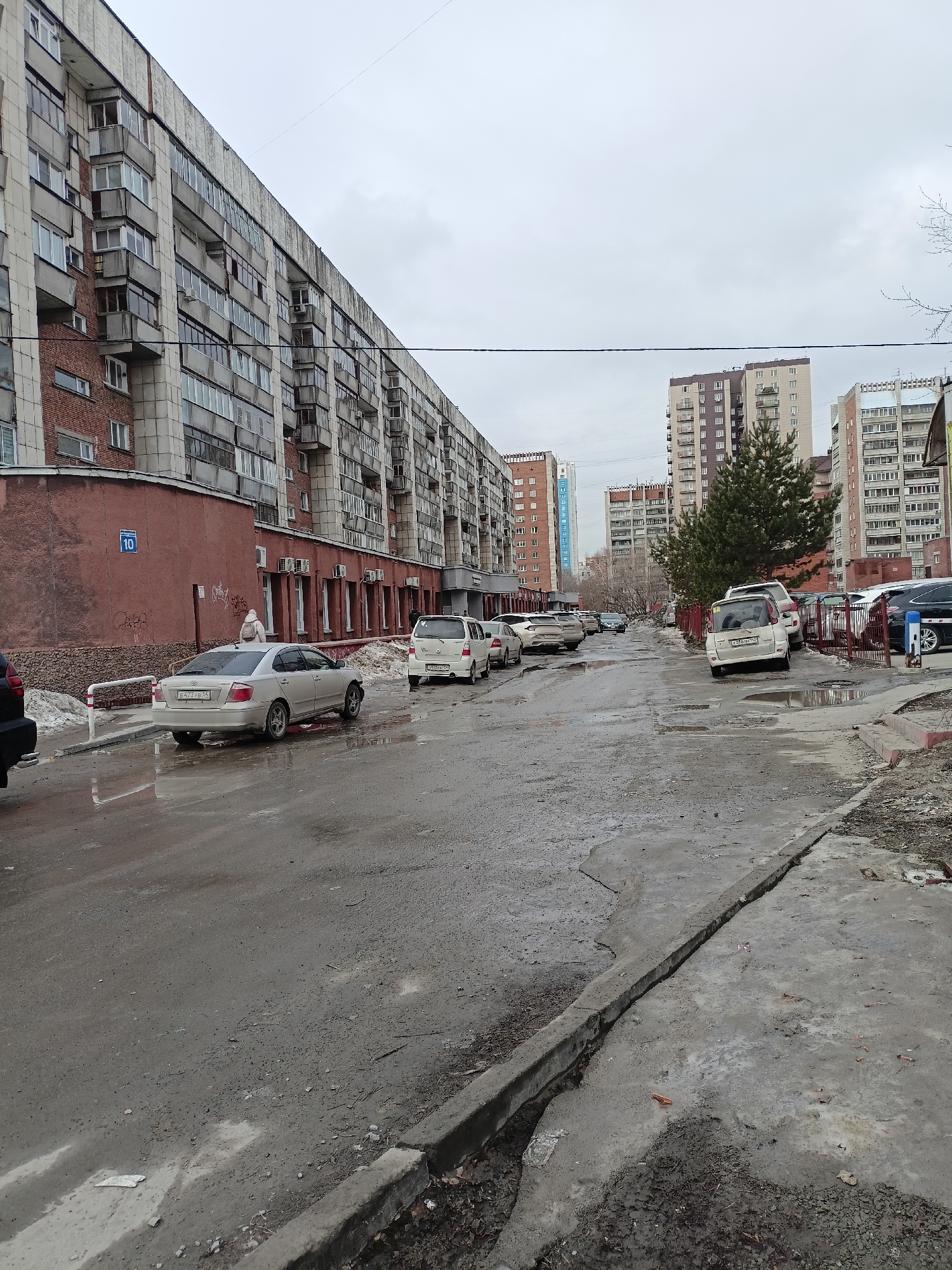 The image depicts a street scene in what appears to be a residential area in Novosibirsk, Russia, based on architectural styles and license plates. The weather is overcast, with wet pavement and remnants of snow visible. A long, multi-story building dominates the left side of the frame, with brick accents and numerous balconies. In the center, the street is pockmarked with potholes and puddles. Several parked cars line both sides of the street. A person wearing a light-colored jacket and hat stands near a building marked with the number "10" (likely a building number). In the background, taller apartment buildings rise above the immediate surroundings. A solitary green tree provides a contrast to the grayness of the day. The overall impression is one of urban decay and a somewhat bleak atmosphere.