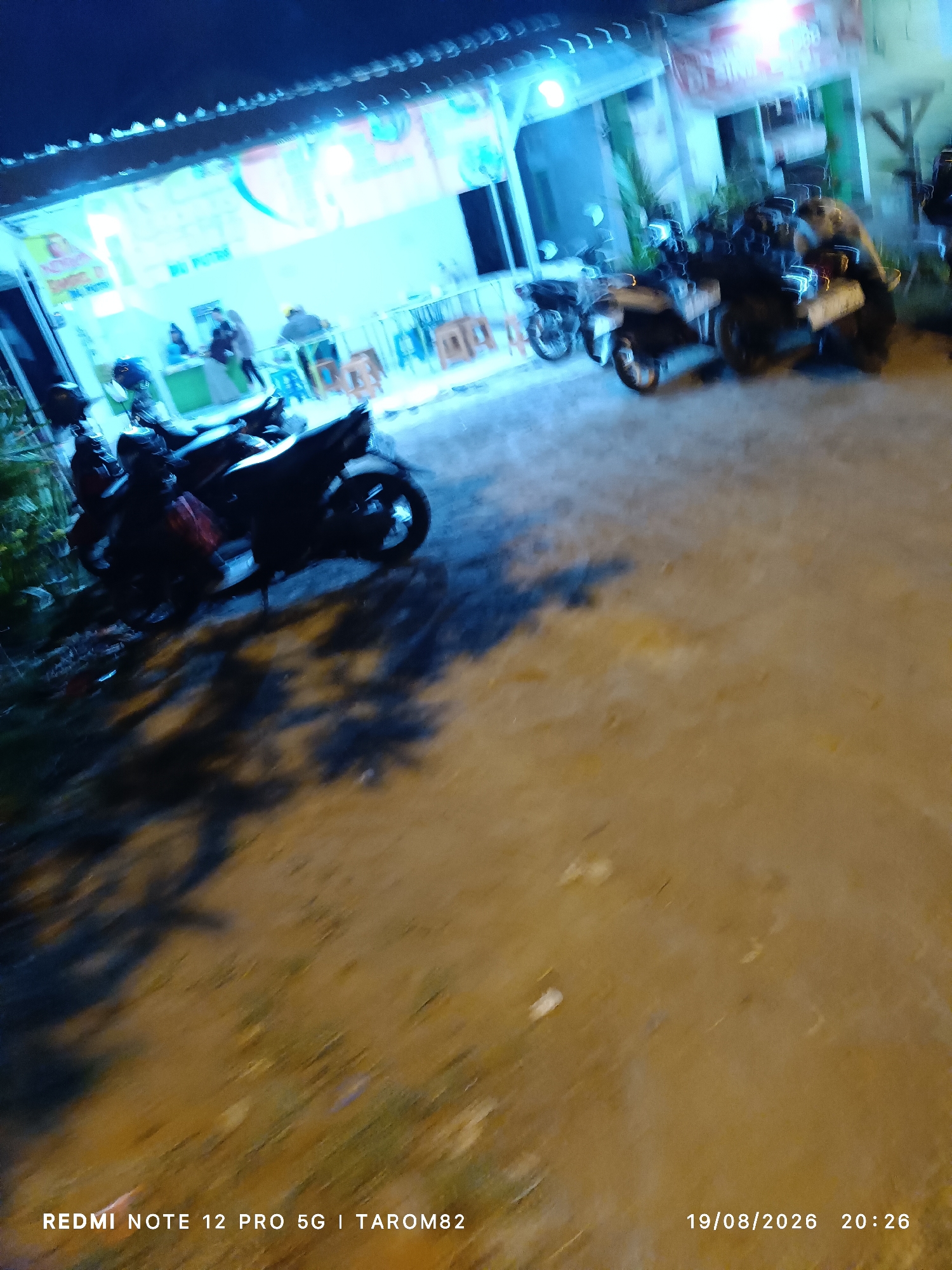 This is a nighttime photograph taken at a street-side establishment in Sidodadi, Indonesia. In the foreground, multiple motorcycles are parked. The middle ground shows a building with bright lighting, likely a restaurant or shop, with several people visible inside and near the entrance. The background is dark with some out-of-focus lights. The ground is paved and appears to be a dirt or gravel surface. The timestamp on the image indicates it was taken on August 19, 2026, at 20:26.