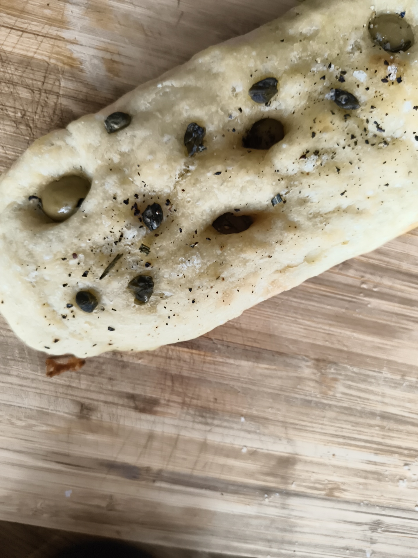 The image shows a piece of focaccia bread on a wooden cutting board. The bread is topped with capers, coarse salt, black pepper, and possibly rosemary. The cutting board shows scratches and wear. The location of the image is not discernible from the visual information provided.