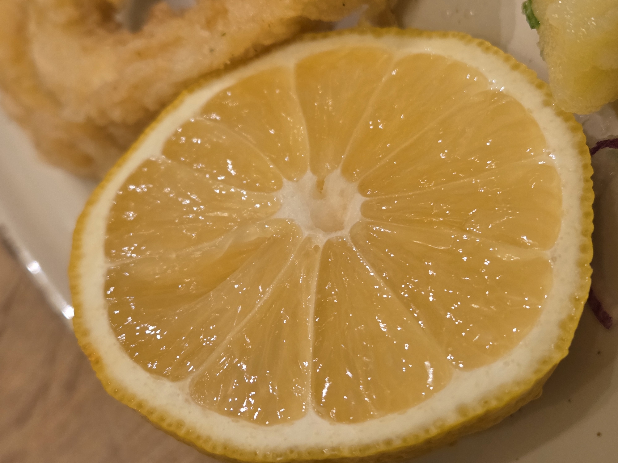 The image shows a close-up of a lemon slice, likely served as a garnish with a meal. Adjacent to the lemon are unidentified fried food items. The scene appears to be at a restaurant, possibly in Varna, Bulgaria, based on the implied context of a prepared meal. However, specific location details in Varna are impossible to determine from the image. The focus is on the details of the lemon slice, emphasizing its cut surface, texture, and juice vesicles.