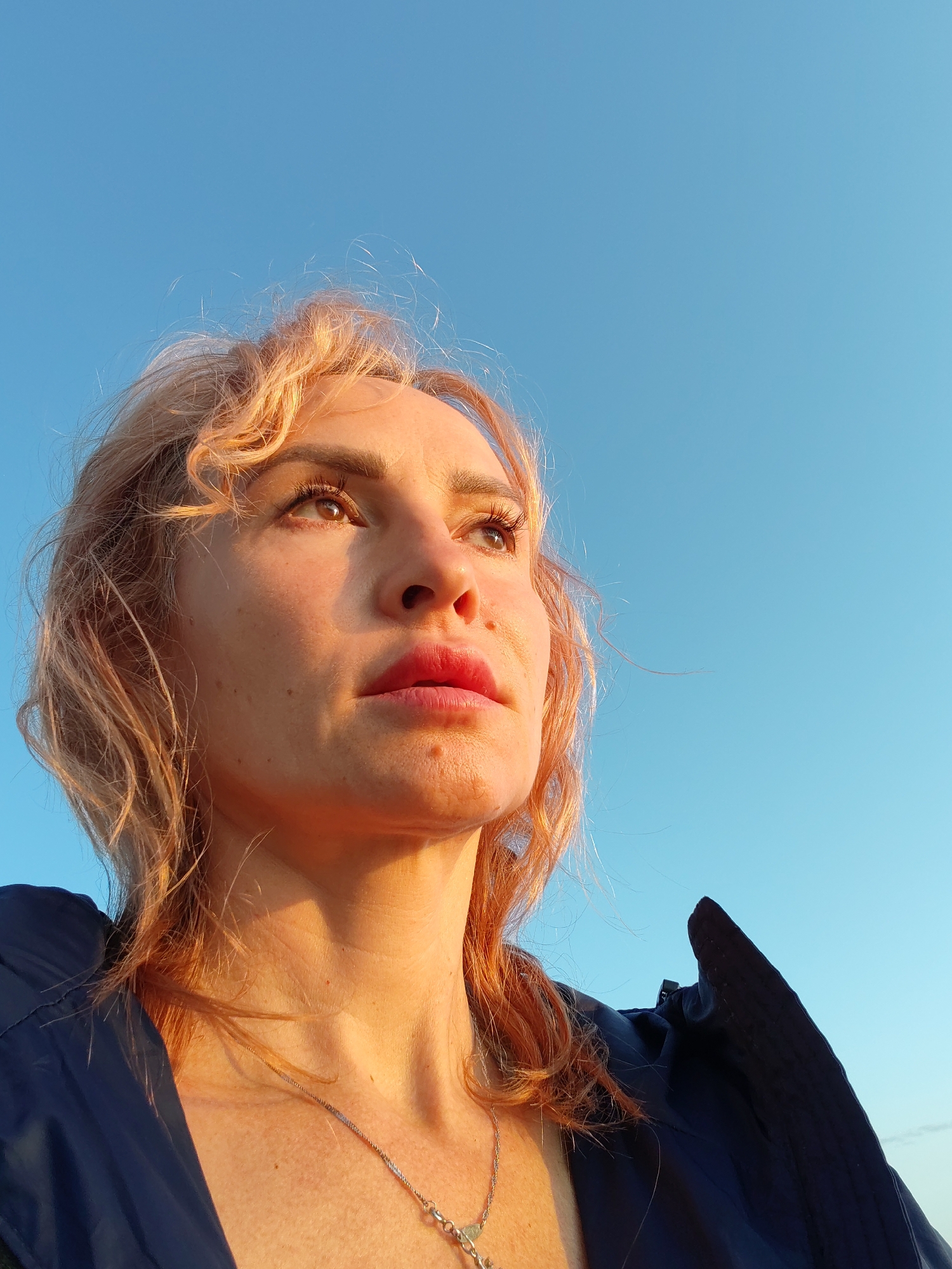 A close-up, low-angle outdoor shot captures the upper body of a woman against a clear, light blue sky. The woman has light auburn, wavy hair that is blowing slightly in the wind. Her head is tilted upwards and to her left, with her gaze directed off-camera. Her skin is illuminated by warm, golden light, highlighting freckles on her cheeks and neck. She has defined eyebrows, visible eyelashes, and full lips. She is wearing a dark blue jacket or coat, and a delicate silver necklace with a pendant is visible around her neck. The background is entirely the sky, with no city elements visible. The image does not provide any information about the location in Kakkarovo, Russia.