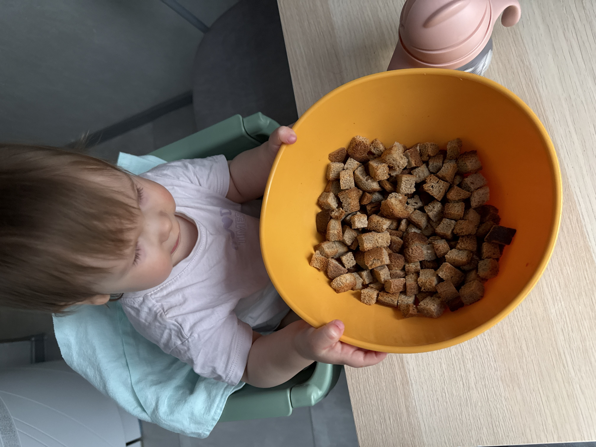 An overhead, high-angle shot captures a young child, likely a baby or toddler, seated in a green high chair at a light wooden table. The scene appears to be indoors, possibly in a kitchen or dining area in Moscow, Russia.
The child has light brown hair and is wearing a white short-sleeved t-shirt, with a light blue fabric (perhaps a bib or blanket) draped over them. Their head is tilted, and their eyes are focused on a large, bright orange bowl positioned directly in front of them. The bowl is filled to the brim with numerous small, cube-shaped pieces of brown croutons or toasted bread.
The child's small hands are visible, gently gripping the rim of the orange bowl. Behind the bowl, a pink sippy cup with a uniquely shaped, perhaps animal-themed, lid is resting on the wooden table. The background is simple, featuring a dark grey wall or furniture behind the child. The lighting suggests it is daytime, and the child's expression appears calm and focused on the food. Part of the word "TIME" is faintly visible on the child's white shirt.