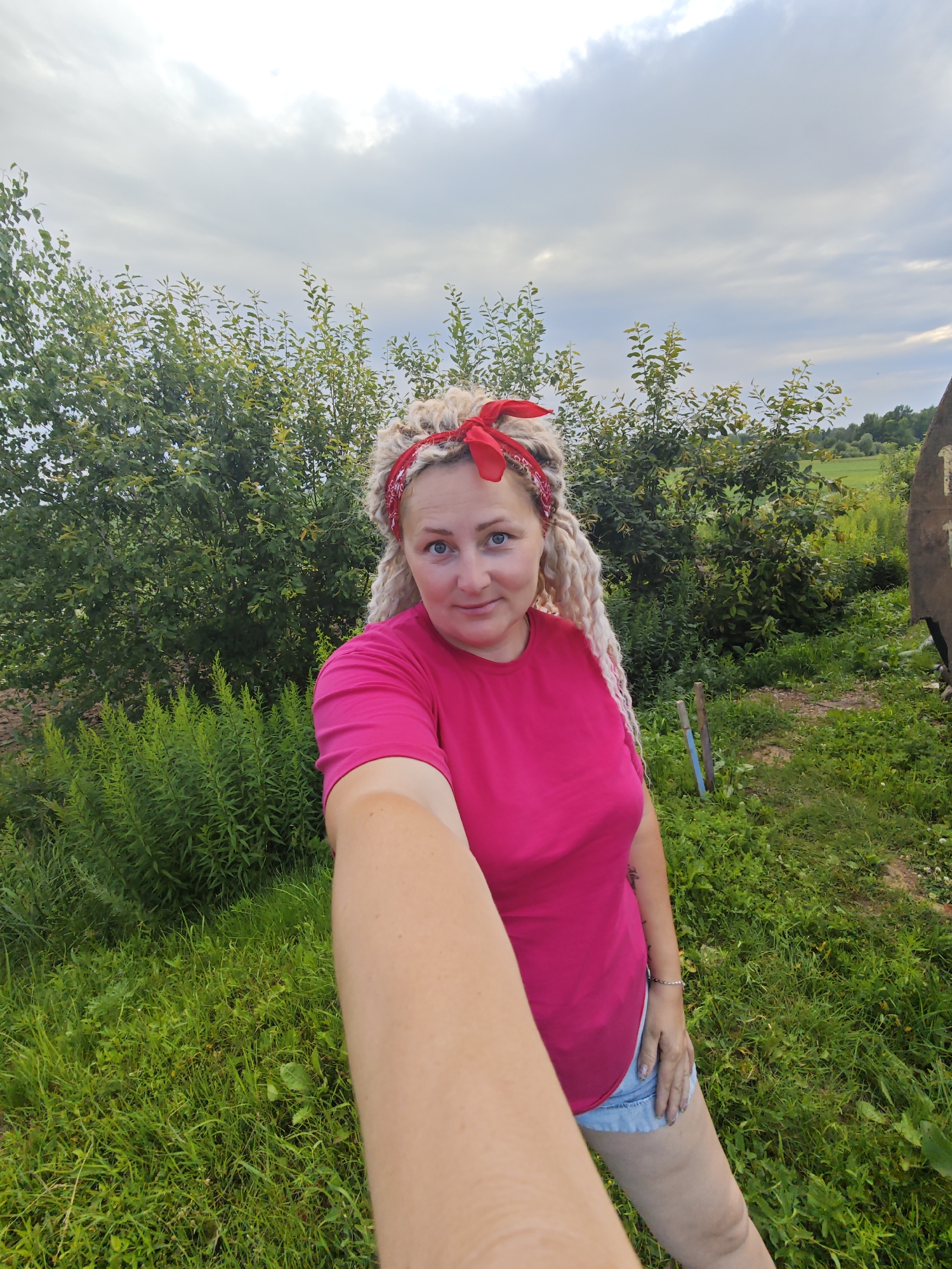 A selfie is taken outdoors in Novoe Mart'yanovo, Russia, on a cloudy day. The subject, a woman with blonde dreadlocks and a red bandana, is wearing a pink t-shirt and denim shorts. She is standing in a grassy area with trees and bushes behind her. There is a wooden object with writing on it to the right of the frame. Two wooden stakes are visible in the grass to the left. The woman is holding the camera with her right arm extended, capturing her upper body and a portion of the surrounding environment.