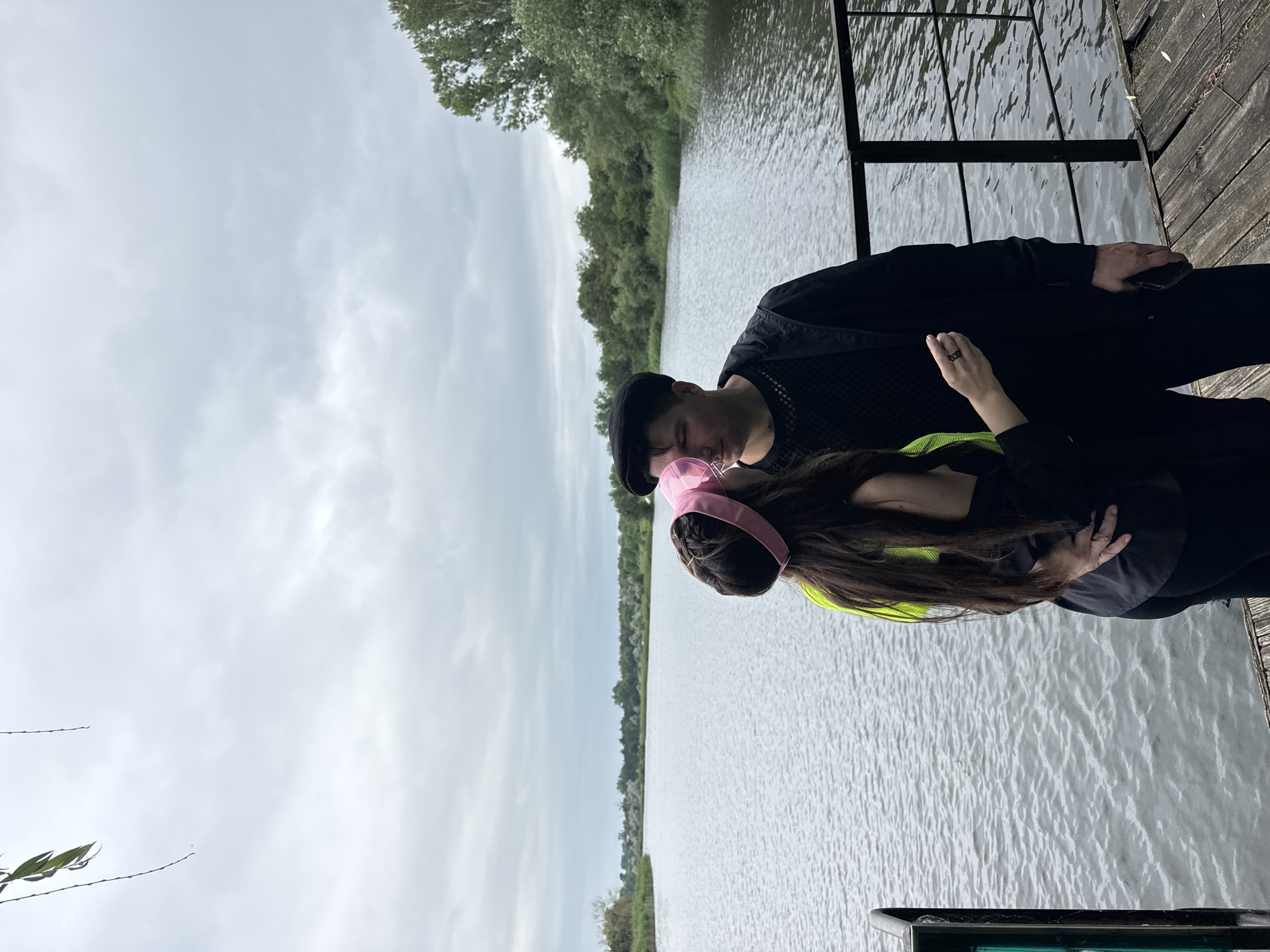 A couple stands on a wooden pier by a body of water. The man, wearing a black beret and jacket, faces the woman, who is wearing a neon yellow top and a pink headband. They are facing each other and appear to be kissing or about to kiss. The water is calm and reflects the overcast sky. Trees and vegetation line the opposite bank of the water. The scene is in Lozuvatka, Ukraine.