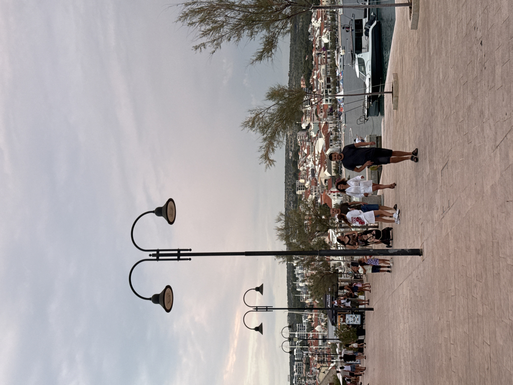The image depicts a promenade in the city of Primošten, Croatia. The promenade is paved with light-colored bricks. In the foreground, two lampposts with double lamps are visible. Several people are walking along the promenade. On the left, a group of people including children are walking, and a woman with a baby in a stroller is also present. Towards the right, a man is walking towards the camera, and a woman is walking away. In the background, a body of water with several boats is visible, and beyond that, the city of Primošten is spread out with its characteristic red-tiled roofs and buildings. The sky is cloudy.