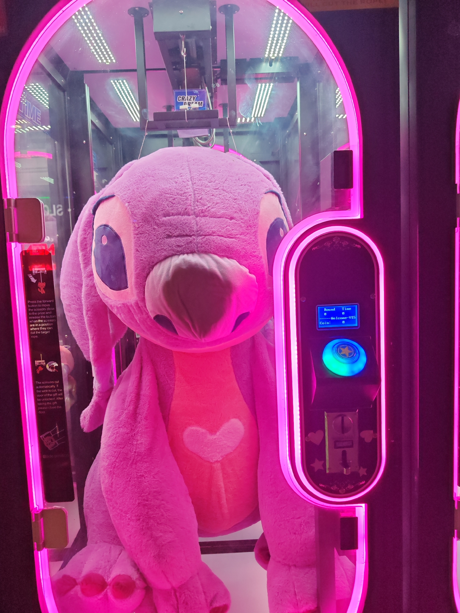 The image shows a pink claw machine with a large, pink stuffed animal resembling a version of Disney's Stitch inside. The machine features a clear glass front illuminated with pink neon lighting, revealing the claw mechanism and the plush toy. A control panel on the right includes a blue illuminated button, a small screen displaying game information, and a coin slot. Instructions are visible on the left side of the machine, explaining how to operate the claw mechanism. The scene is likely from an arcade or entertainment venue. The image does not provide enough information to identify its location in the city of Nesebar, Bulgaria.