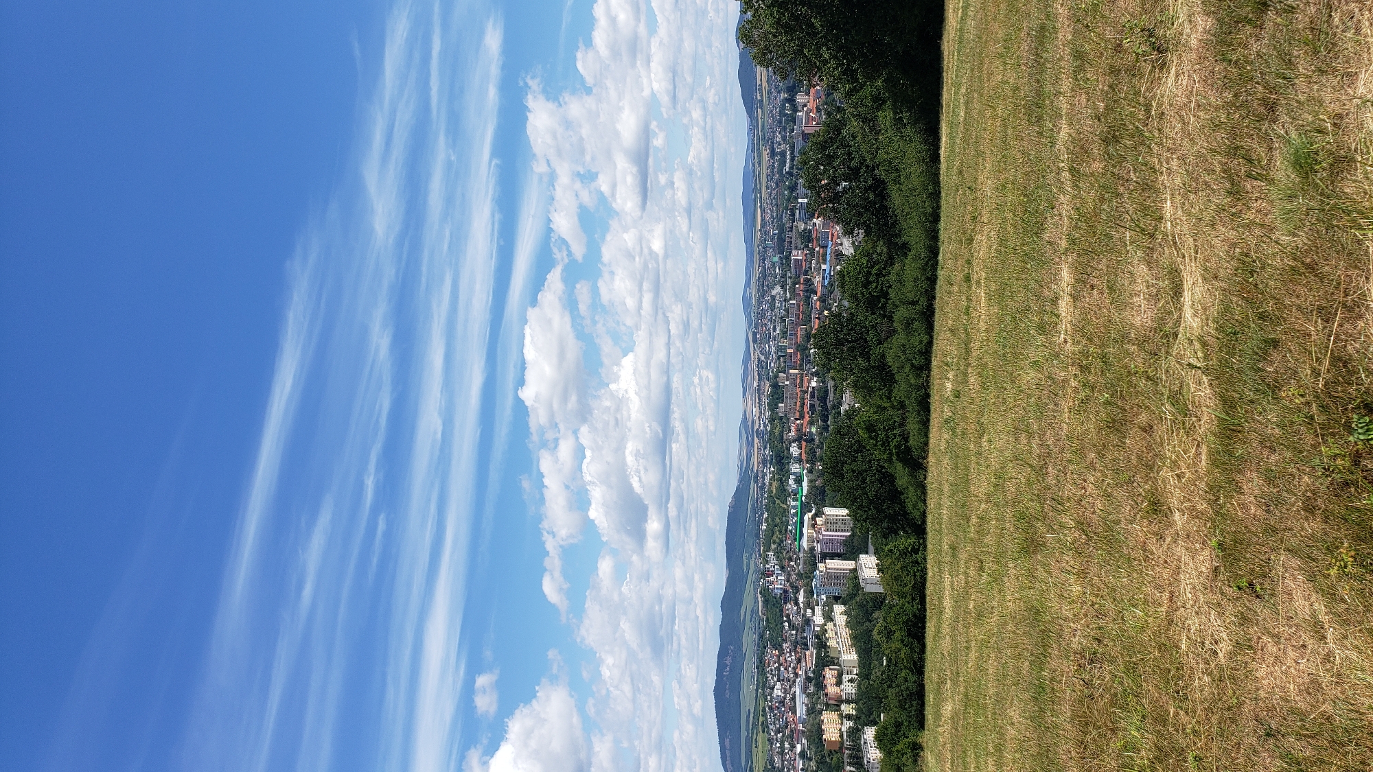 The image captures a panoramic view of the city of Prešov, Slovakia, from a grassy hillside. In the foreground, a sun-drenched field of dry, golden grass slopes gently downwards. Behind the field, a dense line of green trees forms a natural border, partially obscuring the view of the city below.
The city itself is a sprawling urban landscape with a mixture of building types, including residential apartment blocks, some with colorful facades, and lower-rise structures. The buildings are clustered together, creating a dense urban fabric. Beyond the city, a range of low, undulating hills and mountains form the horizon, their silhouettes softened by distance and atmospheric haze.
The sky above is a vibrant blue, dotted with scattered white cumulus clouds. Wispy cirrus clouds streak across the upper portion of the sky, adding texture and depth. The overall impression is one of a clear, bright day, with the city bathed in sunlight. There are no visible people or animals in the image. The scene appears to be a natural landscape overlooking a populated urban area.