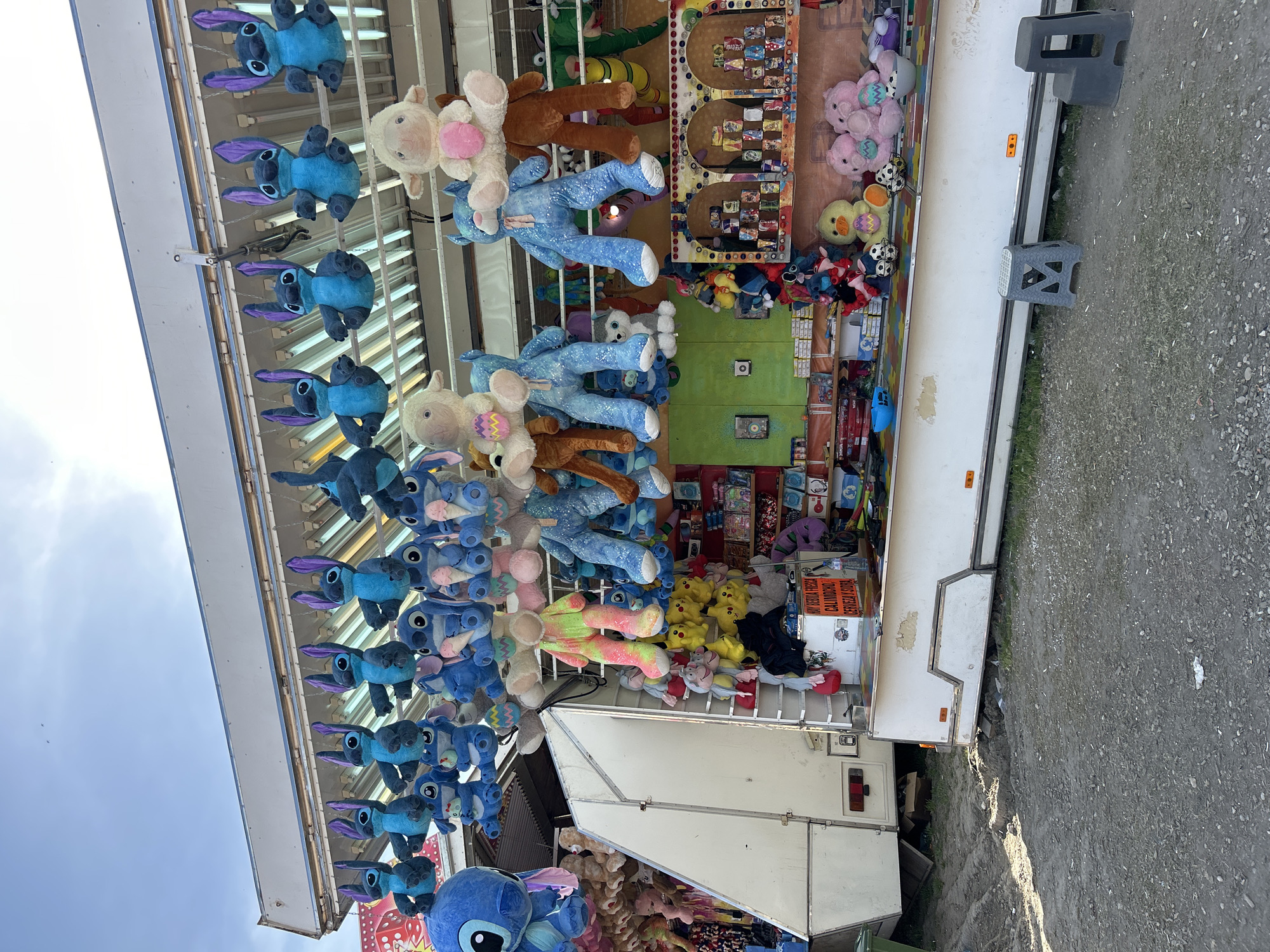 This image captures a vibrant carnival game stall under a bright, partly cloudy sky, likely located at an outdoor fairground in Plovdiv, Bulgaria. The primary subjects are numerous plush toys, prominently displayed as prizes.
A long line of large blue Stitch characters (from Disney's Lilo & Stitch) hangs across the top of the stall, interspersed with several white and pink sheep/lamb plushies. Inside the booth, more stuffed animals are visible, including yellow ducks with Easter egg designs and various other cartoon characters, indicating a wide array of potential winnings.
The stall itself is a white, trailer-like structure with an open front, where a game board featuring multiple prize slots is set up, suggesting a ball toss or similar skill-based activity. Two gray plastic stools are positioned on the dirt and gravel ground directly in front of the booth, awaiting players. The background shows an expansive blue sky with scattered clouds, indicating a clear daytime setting.
A notable detail is a partially visible sign inside the stall with text in what appears to be Bulgarian. Readable words include "ХОТ ДОГ" (Hot Dog), "ПИЦА" (Pizza), "БУРГЕР" (Burger), "ПАЛАЧИНКИ" (Pancakes/Crepes), "СОК" (Juice), "КАФЕ" (Coffee), and "ЦЕНИ" (Prices). This suggests the stall might also offer refreshments or advertise them, or perhaps these refer to different prize tiers or types of games available. No people are visible, leaving the focus solely on the colorful display and game setup.