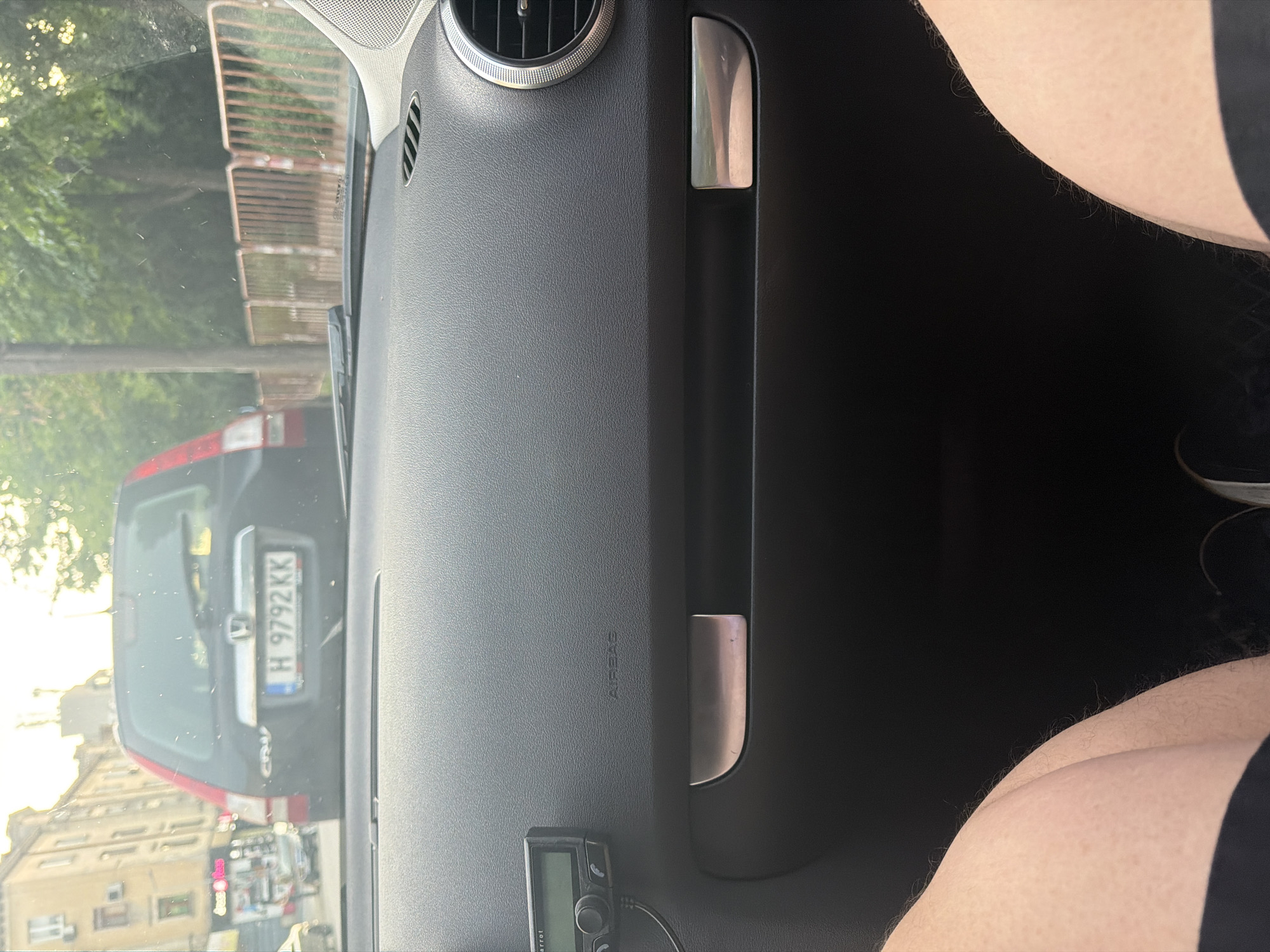 The image shows a view from inside a vehicle, rotated 90 degrees counter-clockwise. The scene captures the car's interior on the left and center, and the exterior through the windshield on the right.
**Subjects:** In the foreground (bottom right of the rotated image), the lower legs or thighs of a person, clad in dark shorts, are visible, suggesting a passenger. The car's dashboard occupies the left and central parts of the image, featuring a circular air vent, a silver passenger grab handle, and an "AIRBAG" inscription. A small black electronic device, branded "Parrot," is attached near the bottom-left of the dashboard. Outside the car (right side of the rotated image), a dark-colored Honda CR-V is prominently visible, seemingly parked directly in front.
**Scene and Setting:** The perspective suggests the photograph was taken from the passenger seat of a stationary car. The exterior view reveals urban or semi-urban surroundings with buildings, green trees, and a brown metal fence. The location is Shumen, Bulgaria.
**Activity or Event:** The primary activity is observation from within a parked or stopped vehicle. No dynamic activity or specific event is captured.
**Notable Details:** The image is brightly lit by natural sunlight, indicating daytime with clear weather. Reflections are visible on the car's interior surfaces and windshield. The license plate on the Honda CR-V, "H 9792 KK," includes the "H" regional code, confirming the location as Shumen, Bulgaria.
**Visible Text:**
* "AIRBAG" on the dashboard.
* "Parrot" on the electronic device.
* "H 9792 KK" on the license plate, with "BG" visible on the blue EU strip.
* "CR-V" on the rear of the Honda.