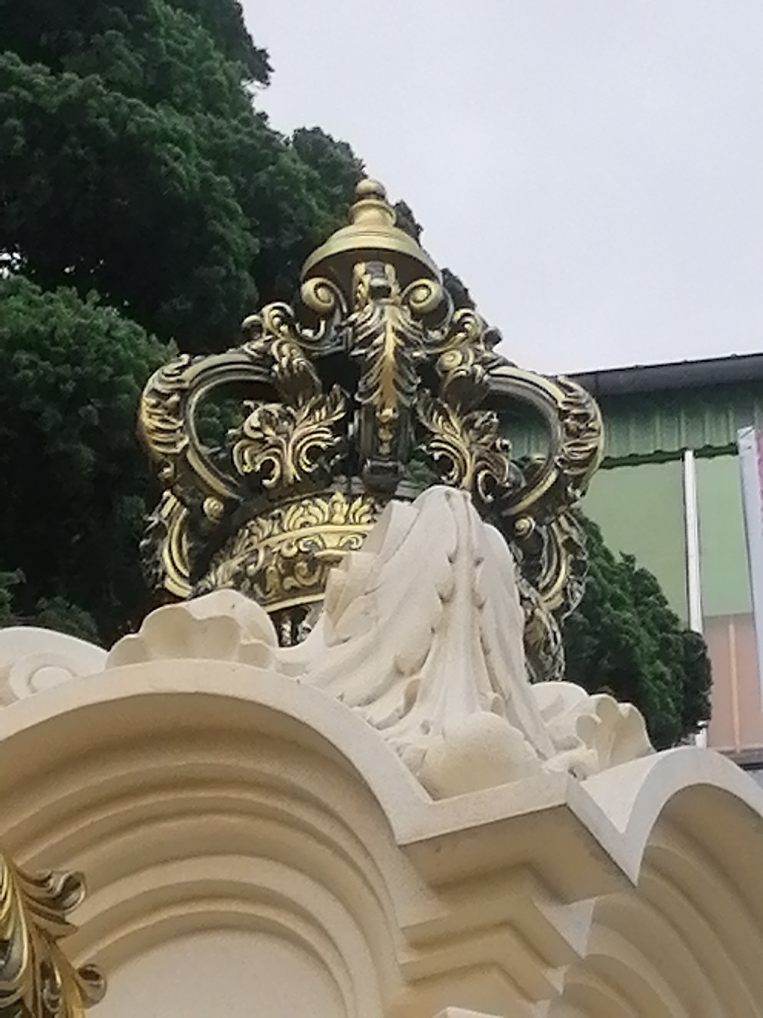 The image captures a large, ornate golden crown sculpture, viewed from a low angle looking upwards. The crown is intricately detailed with abundant scrollwork and leaf-like motifs, suggesting a classical or baroque style, and appears to be crafted from polished metal or a material painted gold. It sits atop a sculpted base, likely made of stone or a similar material, which is light-colored (white or cream) and features curved, layered arches and decorative elements that echo the crown's grandeur.
In the background, dense, dark green foliage from numerous trees creates a lush, natural backdrop on the left and central parts of the frame. To the right, a modern building is partially visible, characterized by a green corrugated metal roof and a large window or glass section with a greenish tint, framed by a vertical white structure.
The scene is lit by soft, diffused light, indicating an overcast sky or a time of day without direct harsh sunlight. There are no people visible, and no specific event or activity is taking place; the focus is entirely on the static sculpture. No text is discernible in the image. The elaborate style of the crown and base, set against the natural greenery and juxtaposed with the modern building, could be a reflection of the diverse architectural and cultural influences found in Da Lat, Vietnam.