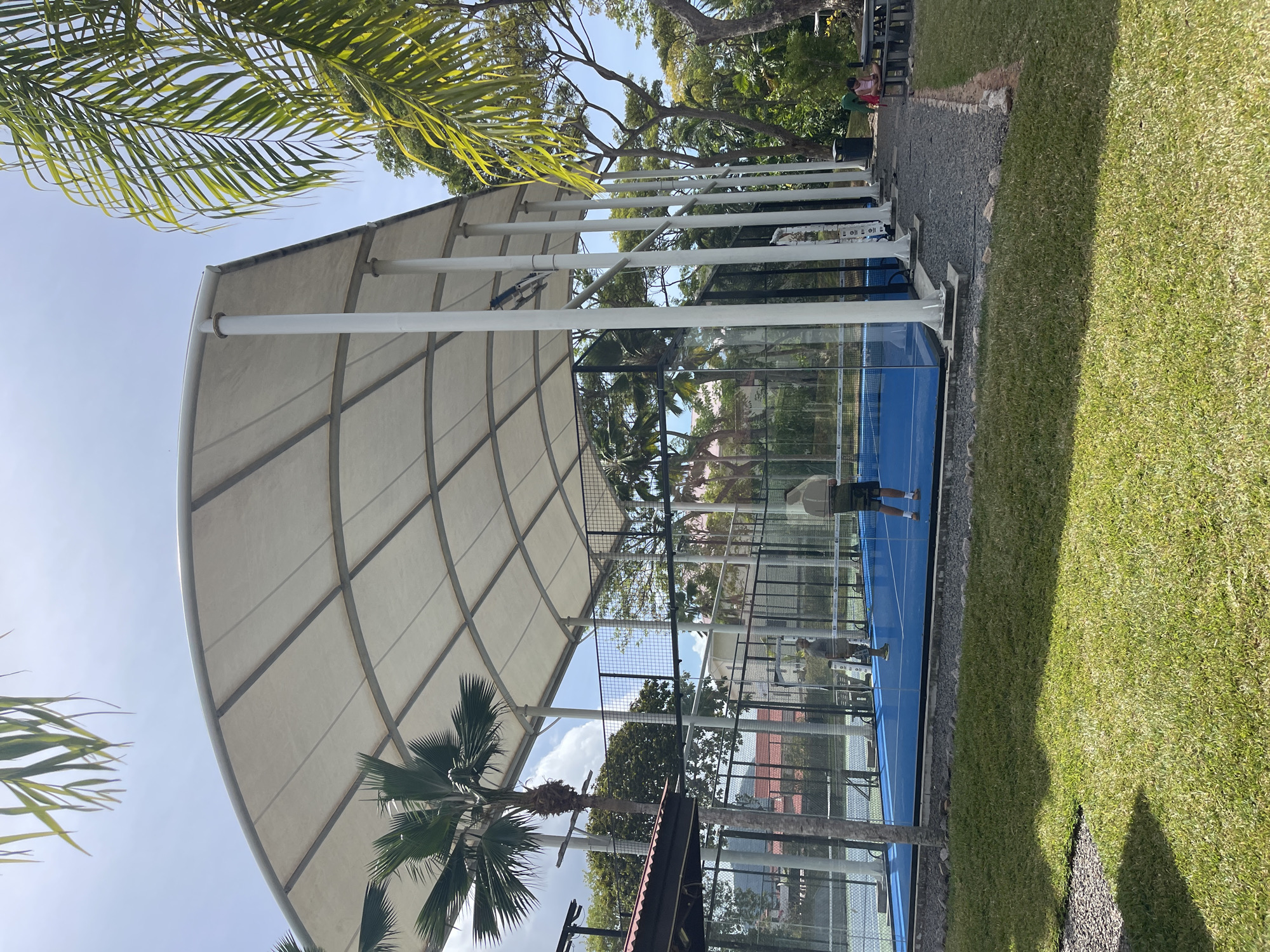 In Providence, Seychelles, a covered outdoor padel tennis court is depicted under a large, white, arched canopy supported by white pillars. The court itself is vibrant blue with white boundary lines. Two individuals are visible on the court, seemingly engaged in a game. One person, wearing a grey t-shirt and shorts, is positioned in the center of the court, while another, dressed in a white long-sleeved top and dark shorts, stands near the net. The court is enclosed by glass walls and black mesh fencing, typical of padel courts. Lush green grass surrounds the court area, and several palm trees are scattered throughout the landscape, indicating a tropical environment. In the background, more trees and buildings can be seen, suggesting a sports complex or recreational area. A young person is seated at a picnic table to the right of the court, observing the game. The sky is clear with a few scattered clouds.