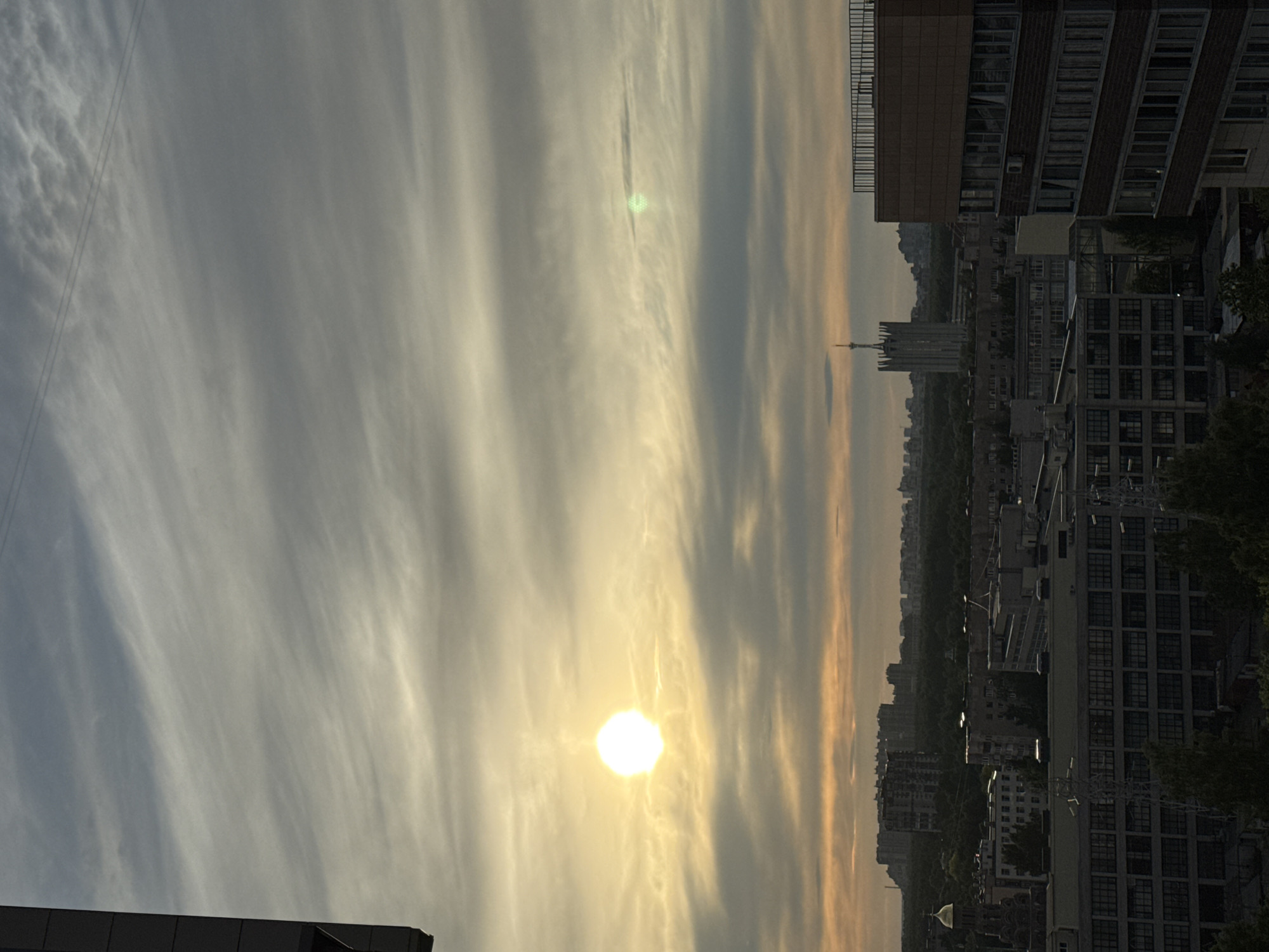 This image captures a panoramic view of a city skyline under a dramatic sky, likely at sunrise or sunset. The sky is dominated by long, wispy, horizontally stretched clouds, suggesting high-altitude cirrus formations, which are illuminated by the low-lying sun. The sun itself appears as a brilliant, intense white orb positioned towards the right side of the frame, casting a warm, golden hue across the clouds and creating a bright lens flare.
In the mid-ground and background, a sprawling urban landscape unfolds. Numerous buildings, many with repeating window patterns, are silhouetted against the sky, their details obscured by distance and the low light. A distinctive, tall, spired building, resembling a Gothic-style skyscraper or a prominent tower, stands out in the central-right portion of the city's horizon, drawing the eye. Dark patches of trees or foliage are interspersed among the buildings, particularly on the right side of the frame. Two faint, parallel power lines are visible against the cloudy sky in the upper left quadrant.
The scene conveys a sense of calm and vastness, typical of dawn or dusk. No human activity or visible text is present. The architectural style, particularly the prominent spired structure, suggests a large city, potentially in a European context.