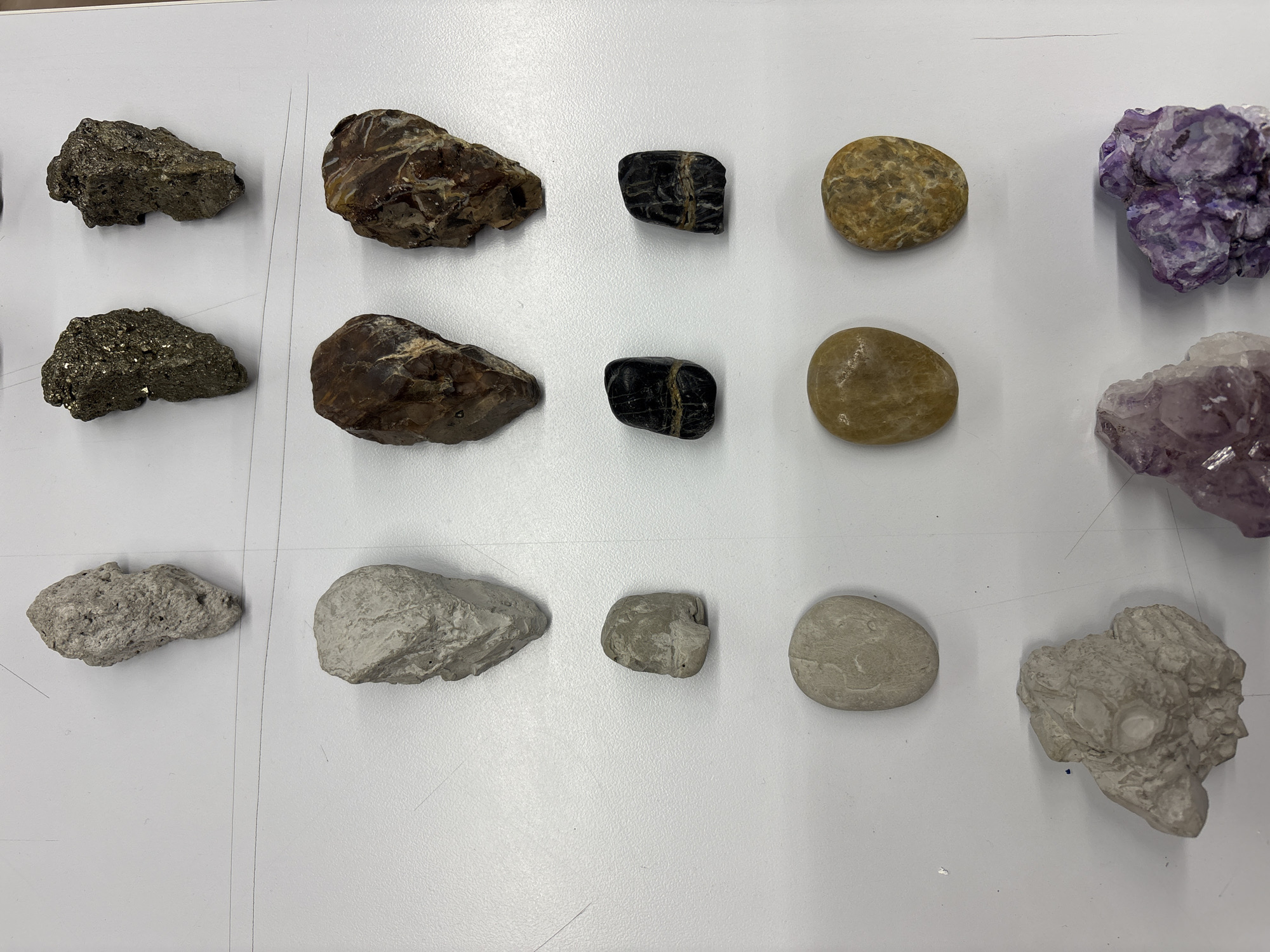 The image shows a collection of rocks and minerals laid out on a white surface. There are 15 rocks and minerals in total, arranged in 3 rows and 5 columns. The materials include pyrite, chert, obsidian, quartz pebbles, amethyst and possible tufa. The setting is indoors with bright artificial lighting. The picture was taken in Mexico City.