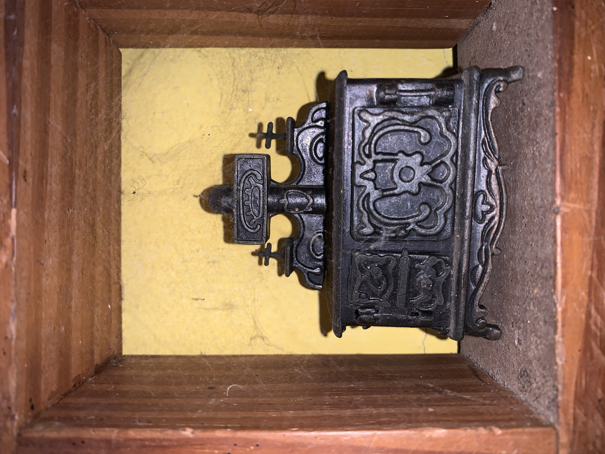 The image shows an antique iron lock mechanism mounted within a wooden frame. The lock is intricately designed with ornate details and is mounted on a yellow wall. The frame appears old, with visible wear, dust, and cobwebs. The overall lighting is dim. The location is unknown.