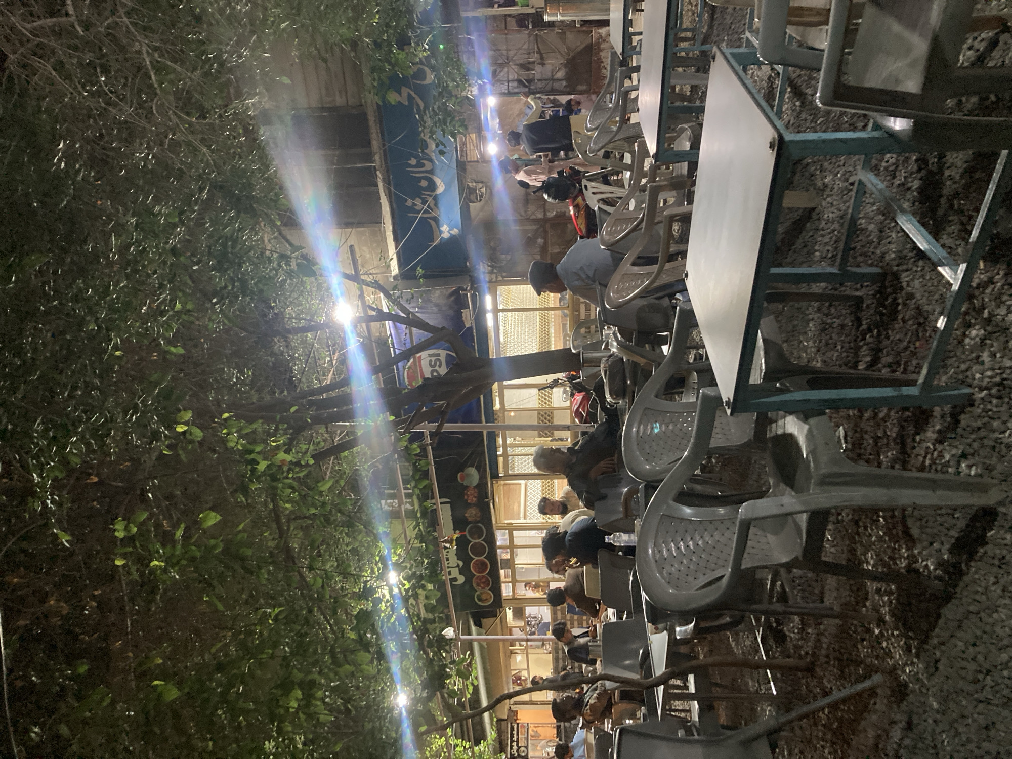 The image depicts an outdoor dining area in Quetta City Tehsil, Pakistan at night. Several people are seated at tables, appearing to be eating or socializing. The tables and chairs are simple, likely made of plastic or metal. The environment includes trees to the left side of the picture and gravel on the ground. A sign or menu board is visible in the background.