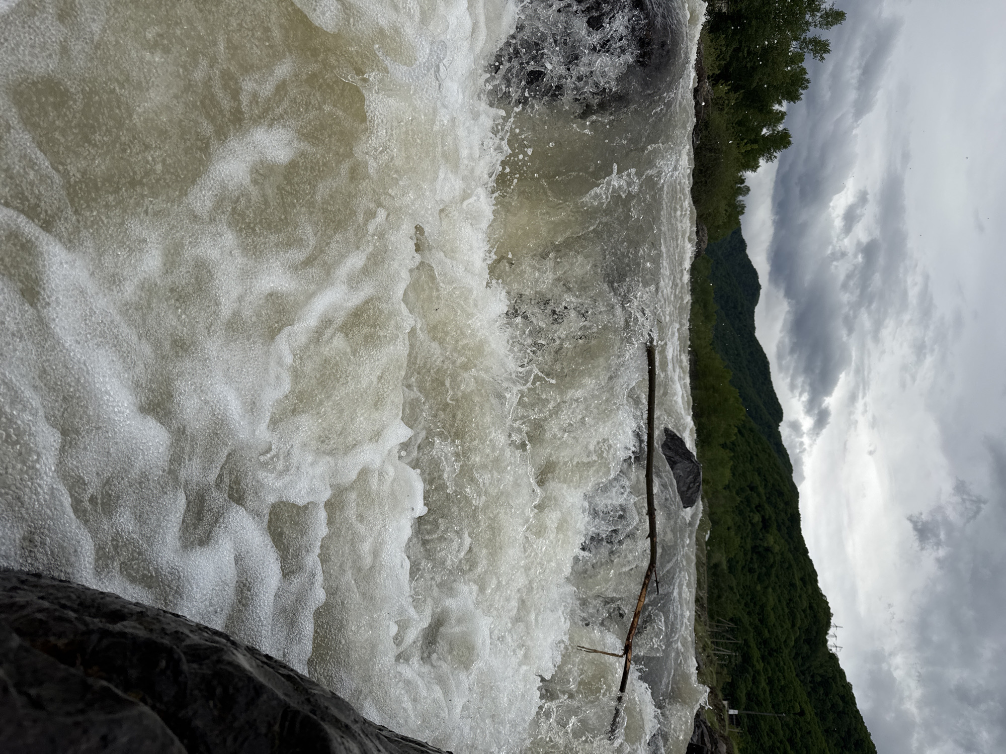 The image presents a close-up, slightly angled view of turbulent water cascading over rocks, with elements of the surrounding landscape visible in the background. The foreground is dominated by the rushing, foamy water, the light, milky color and bubbly texture indicating a fast-moving flow. Dark rocks are visible at the edge of the frame, partially submerged in the water. A thin branch or stick is caught in the current, adding a linear element to the composition. In the background, a tree-lined area and a mountain slope covered with dense green vegetation are visible. The sky is overcast, with grey clouds filling most of the upper portion of the image. Given the location information (Каменномостский, Russia), the scene likely depicts a small waterfall or rapids within a river or stream in the area. The presence of a mountain suggests a geographical context of a mountainous or hilly region.