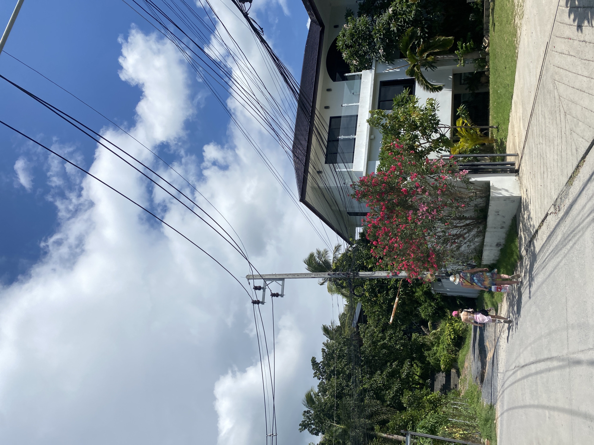 The image depicts a residential street scene in Baan Lamai, Thailand, under a clear blue sky with scattered white cumulus clouds. The foreground features a light grey asphalt or concrete road with faint tire marks. Two individuals, identified as women, are walking away from the viewer along the right side of the road.
The woman on the left wears a light-colored cap, a light pink strapless top, and a matching light pink skirt or sarong, and carries a dark bag on her left shoulder. The woman on the right wears a multi-colored, patterned dress, a light straw hat, and carries a red and white bag in her left hand.
To the left of the road, a dirt and gravel path slopes upwards into dense tropical foliage. A white and orange striped barrier arm is visible near the path entrance. A tall, light-colored concrete utility pole stands prominently on the left, supporting multiple black overhead power lines that traverse the upper portion of the frame from left to right.
On the right side of the road, a well-maintained, two-story white building with a dark-tiled, pitched roof is visible. The building features multiple large, dark-paned windows and a balcony with clear glass railings on the upper floor. In front of the building's perimeter wall, a dense, vibrant pink flowering bush, possibly an oleander or bougainvillea, is in full bloom. A small, dark-colored gate is partially visible next to the bush. Lush green tropical vegetation, including palm trees and various broadleaf trees, surrounds the property and extends into the background.