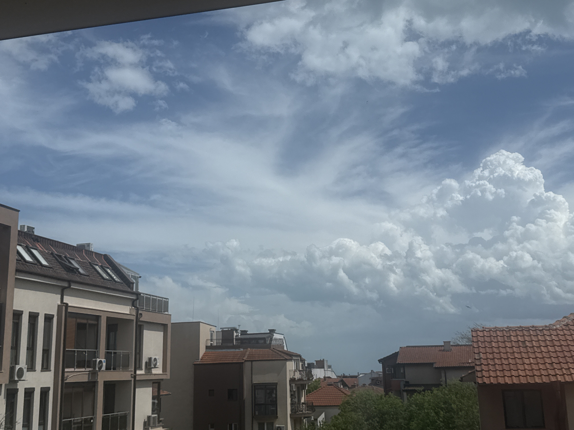 The image depicts a daytime view over a section of Burgas, Bulgaria. The foreground is dominated by buildings with varying architectural styles, including a modern apartment building on the left with multiple balconies and a terracotta-tiled roof structure. Other buildings with red-tiled roofs are also visible. In the background, the sky is filled with a mixture of cirrus and cumulus clouds, suggesting potential changing weather conditions. The overall environment appears to be residential with trees visible.