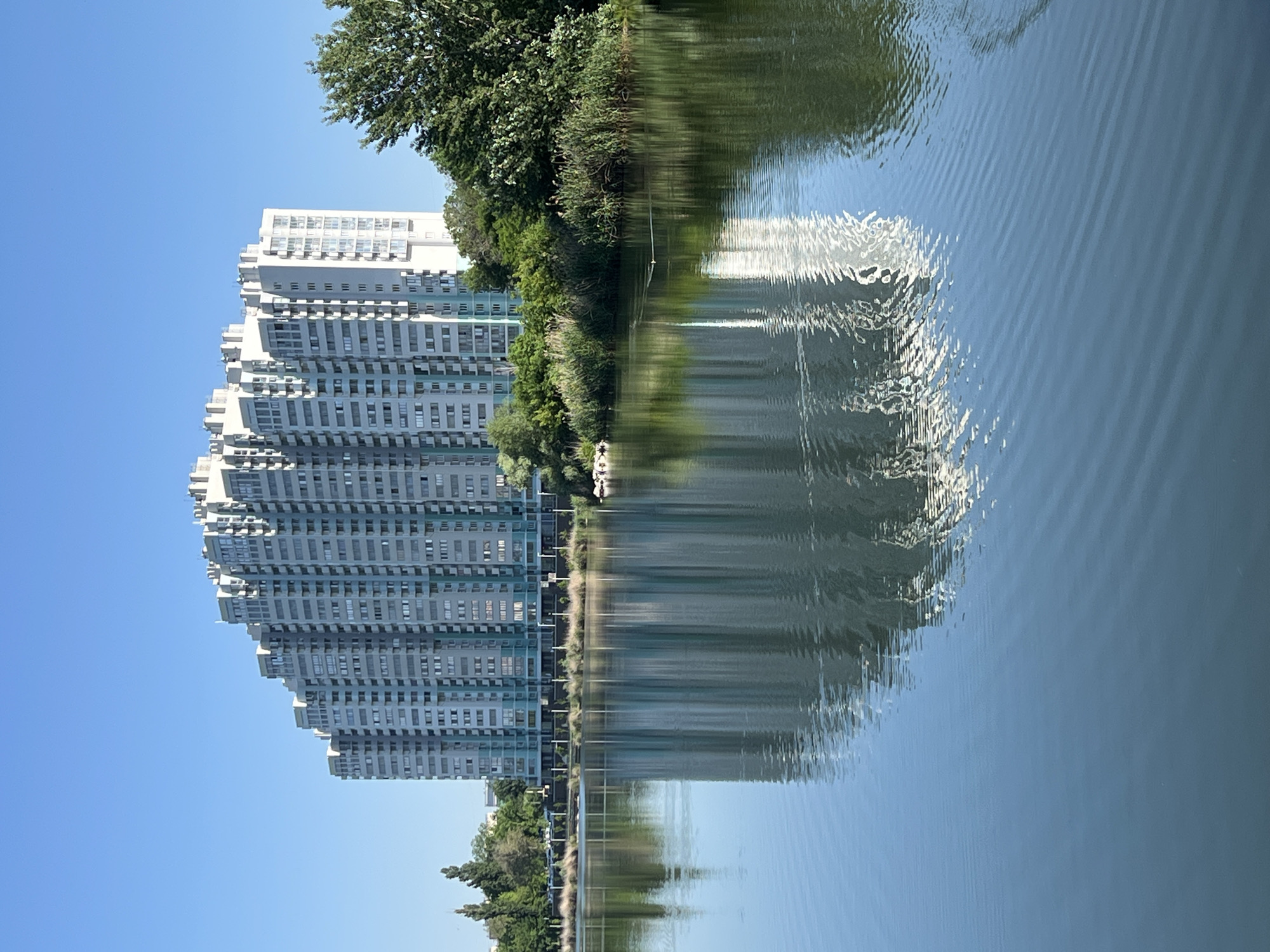 This full-shot, eye-level photograph depicts a large, modern multi-story building prominently reflected in a calm body of water under a clear blue sky. The building, situated in the city of Krasnodar, Russia, exhibits a distinct stepped or terraced architectural profile, rendered primarily in light grey and white, with noticeable light blue/teal accents on its facades and balcony structures. Numerous windows are distributed across its many floors. The structure's reflection occupies the lower two-thirds of the image, appearing with minor distortions caused by concentric ripples on the water's deep blue-green surface. A dense, verdant band of trees, reeds, and bushes delineates the far bank, positioned between the water's edge and the building. The sky above the building is uniformly clear and light blue. No individuals are discernible. A small, light-colored object with a faint wake is visible on the water's surface towards the mid-right, contributing to localized ripples. Additional small white objects are present on the left bank at the waterline.