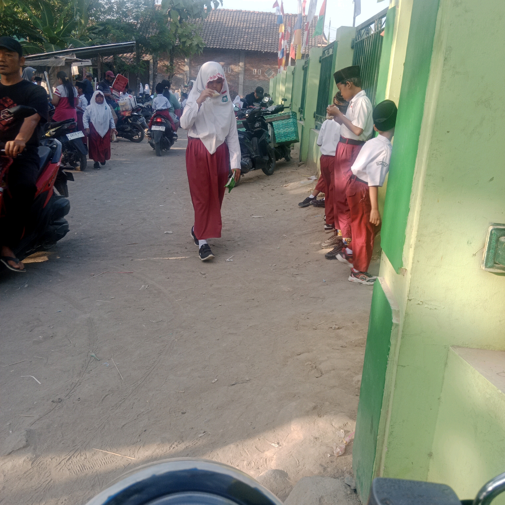 In Tegowanu Kulon, Indonesia, a group of children in school uniforms stand in a line by a green wall. A girl in a white hijab and red skirt walks towards the viewer, eating something from a wrapper. In the background, more children and adults are visible, some on motorcycles, and a building with a tiled roof. The ground is sandy and appears to be a dirt road. Flags are strung up in the distance.