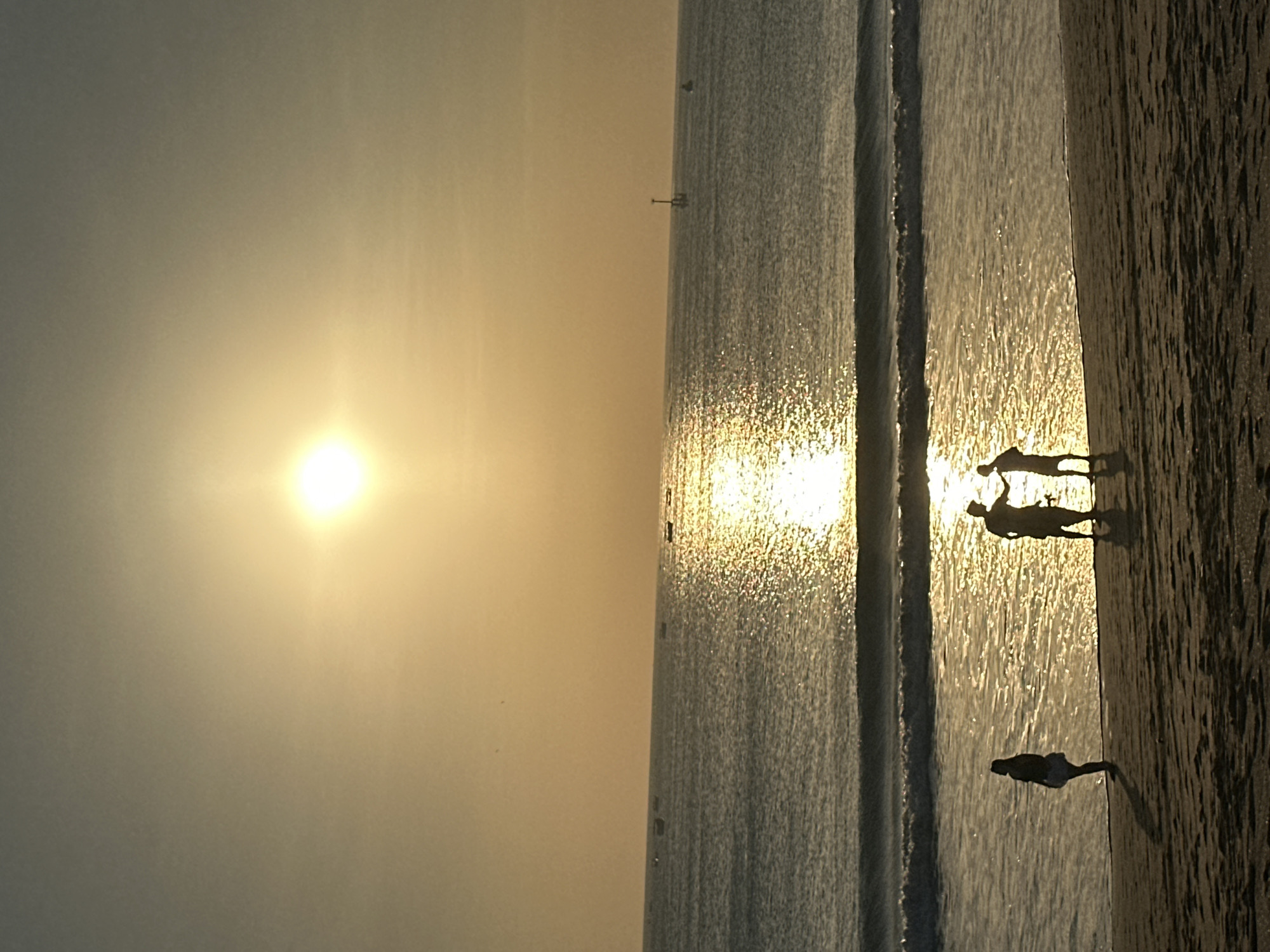 The image depicts a beach scene at sunset in Kuta, Indonesia. The sun is low in the sky, casting a warm glow over the ocean and the beach. Silhouettes of people are visible on the shore. In the foreground, a lone figure stands facing the water. Further along the beach, two figures are interacting, their silhouettes suggesting conversation or a greeting. The ocean has gentle waves, and the water reflects the sunlight, creating a shimmering effect. Several small boats are scattered in the distance on the horizon, and a navigational marker is visible. The sky is a soft gradient of yellow and gray, typical of a sunset. The sand on the beach is dark and textured, with shadows cast by the setting sun.
