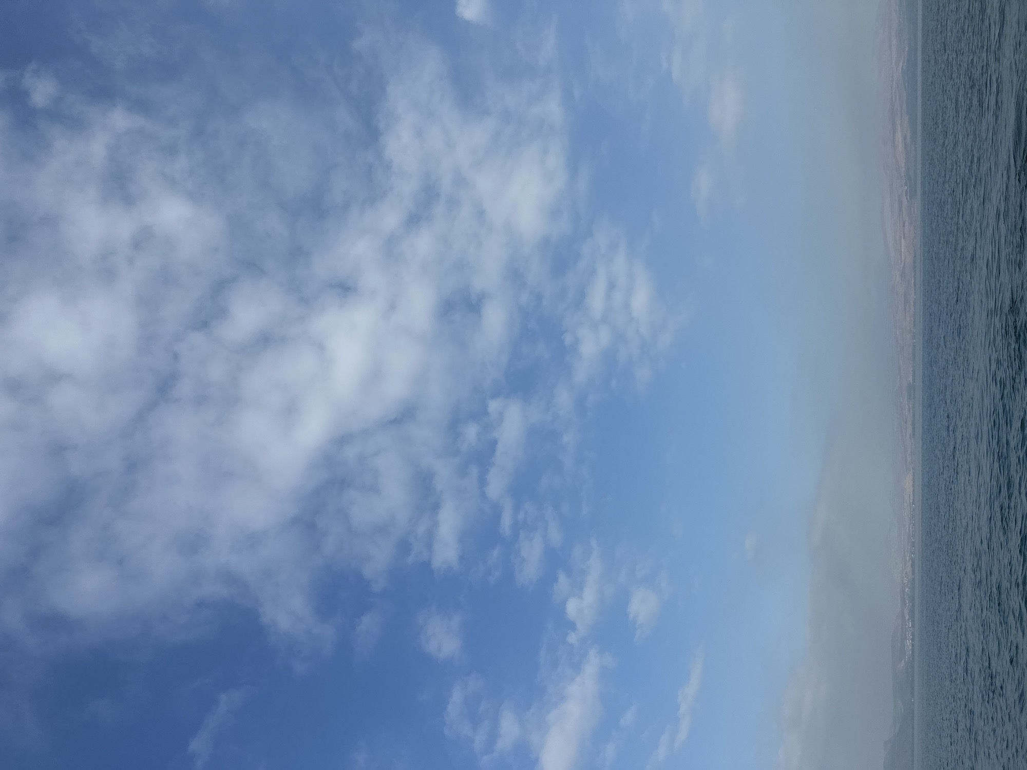 The image depicts a wide-angle view of the Sea of Galilee, with the city of Tiberias, Israel, faintly visible on the distant shore. The sky is a vibrant blue, dotted with scattered white clouds that appear wispy and spread out. The sea occupies the lower portion of the frame, its surface textured with gentle ripples and a deep blue hue. The horizon line is clearly defined, separating the water from the land. The distant shoreline is obscured by a light haze, making the details of Tiberias appear softened and distant. The overall atmosphere is calm and serene, suggesting a clear day with good visibility.