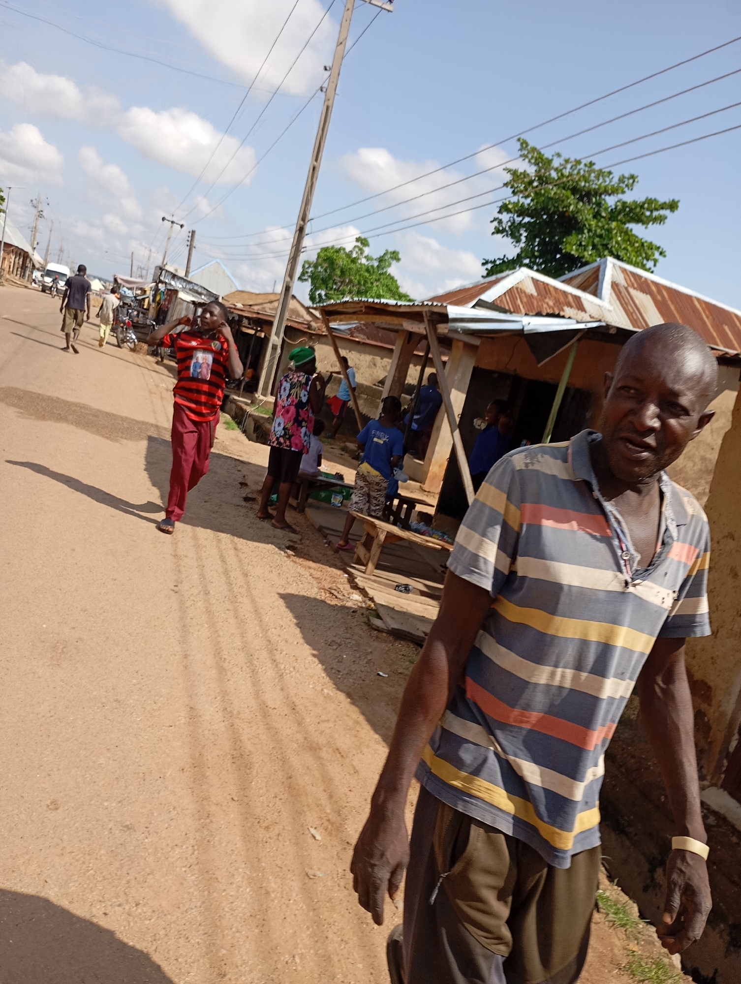 The image captures a street scene in what appears to be a rural or semi-urban area. The time of day seems to be daytime, with bright sunlight casting long shadows on the dusty road. The weather is clear, with a blue sky dotted with white clouds.
In the foreground, a man is facing the camera, looking slightly to the right. He is wearing a horizontally striped t-shirt with colors of blue, gray, yellow, and coral, and dark pants. He has a light-colored bracelet on his right wrist. Behind him, to the left, another man is walking away from the camera, wearing a red and black striped t-shirt and red trousers. Further in the background, several other people are visible, some walking and others standing near small market stalls. Children are also present, with one child wearing a blue t-shirt.
The setting includes several simple structures with corrugated metal roofs, suggesting market stalls or small shops lining the street. Power lines are visible overhead, supported by wooden poles. The road is unpaved and appears to be made of dirt or gravel. The overall atmosphere is one of everyday activity in a community. No text is clearly visible in the image.