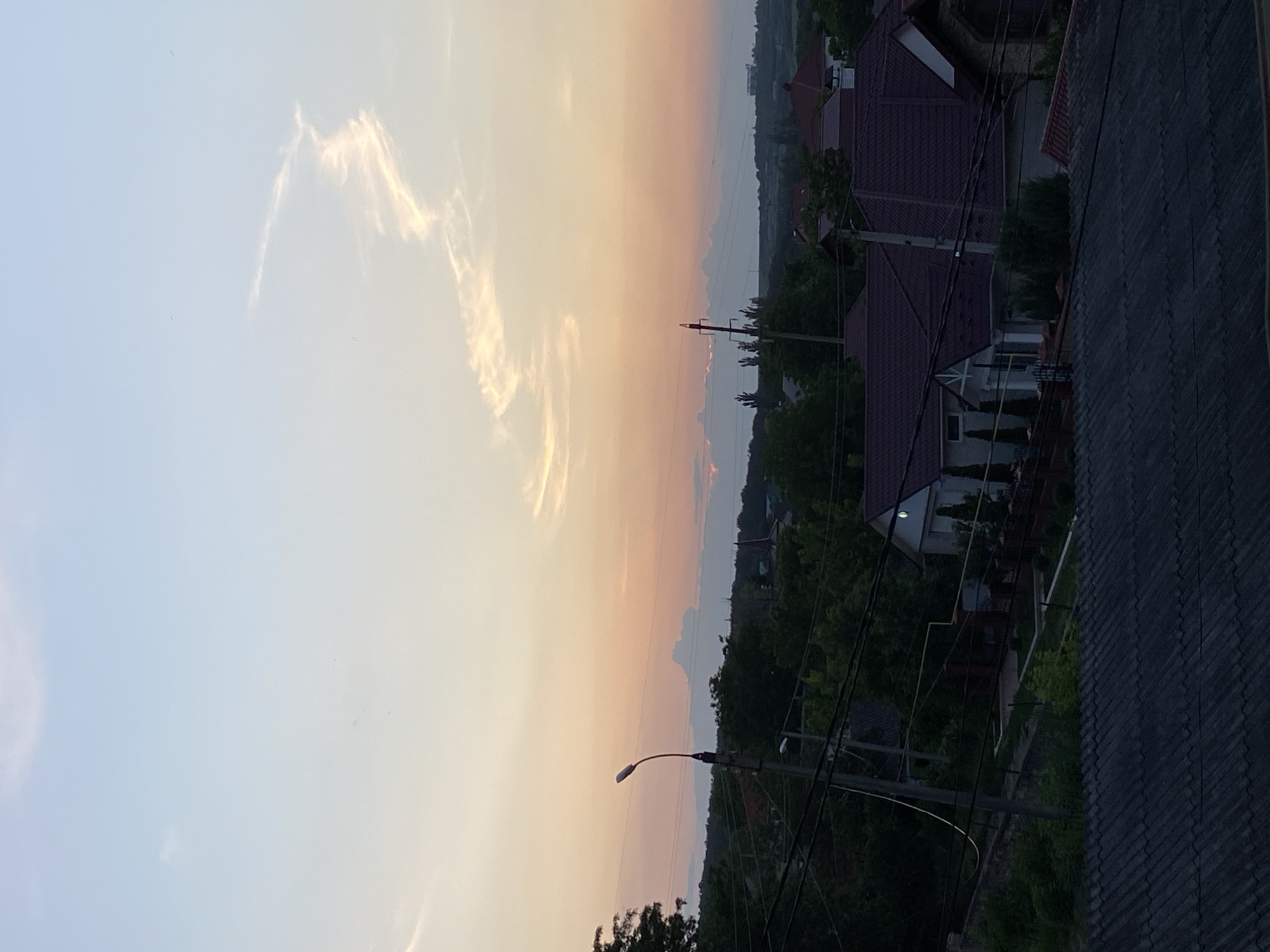 A low-angle shot captures a dusky sky with soft hues of peach and light blue. A wispy, elongated cloud formation snakes across the upper right quadrant of the frame, catching the light of what appears to be a setting sun. Below the sky, a suburban landscape unfolds. In the mid-ground, several houses with dark red tiled roofs are nestled among lush green trees. A solitary streetlamp stands prominently on the left, its light emanating from a single, upward-facing bulb. Power lines crisscross the scene, strung between utility poles. The foreground is dominated by the corrugated texture of a dark, weathered roof, likely a building within the frame. The overall impression is one of a quiet evening in a residential area, with the natural beauty of the sky providing a dramatic backdrop.