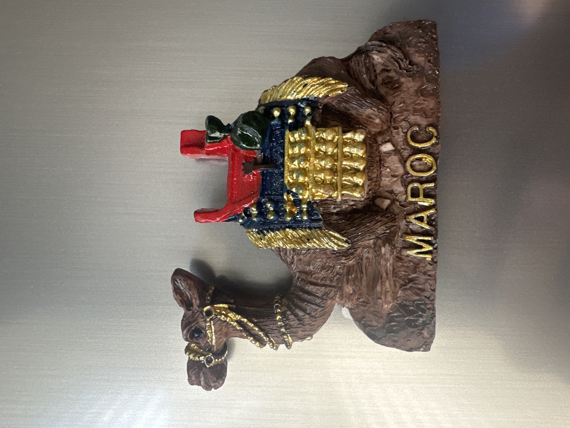 A magnetic souvenir of a camel is attached to a metallic surface, possibly a refrigerator. The camel is depicted in a resting position with its legs folded underneath. It is adorned with a decorative saddle and bridle, both featuring intricate gold accents and what appear to be blue and red fabric details. The camel's base is textured to resemble a rocky or sandy terrain and has the word "MAROC" inscribed in gold lettering. The metallic surface behind the magnet is plain and reflects light, creating a subtle gradient. The scene is well-lit, highlighting the details of the magnet. The location in the city of Ruse, Bulgaria is not depicted or suggested by the image itself.