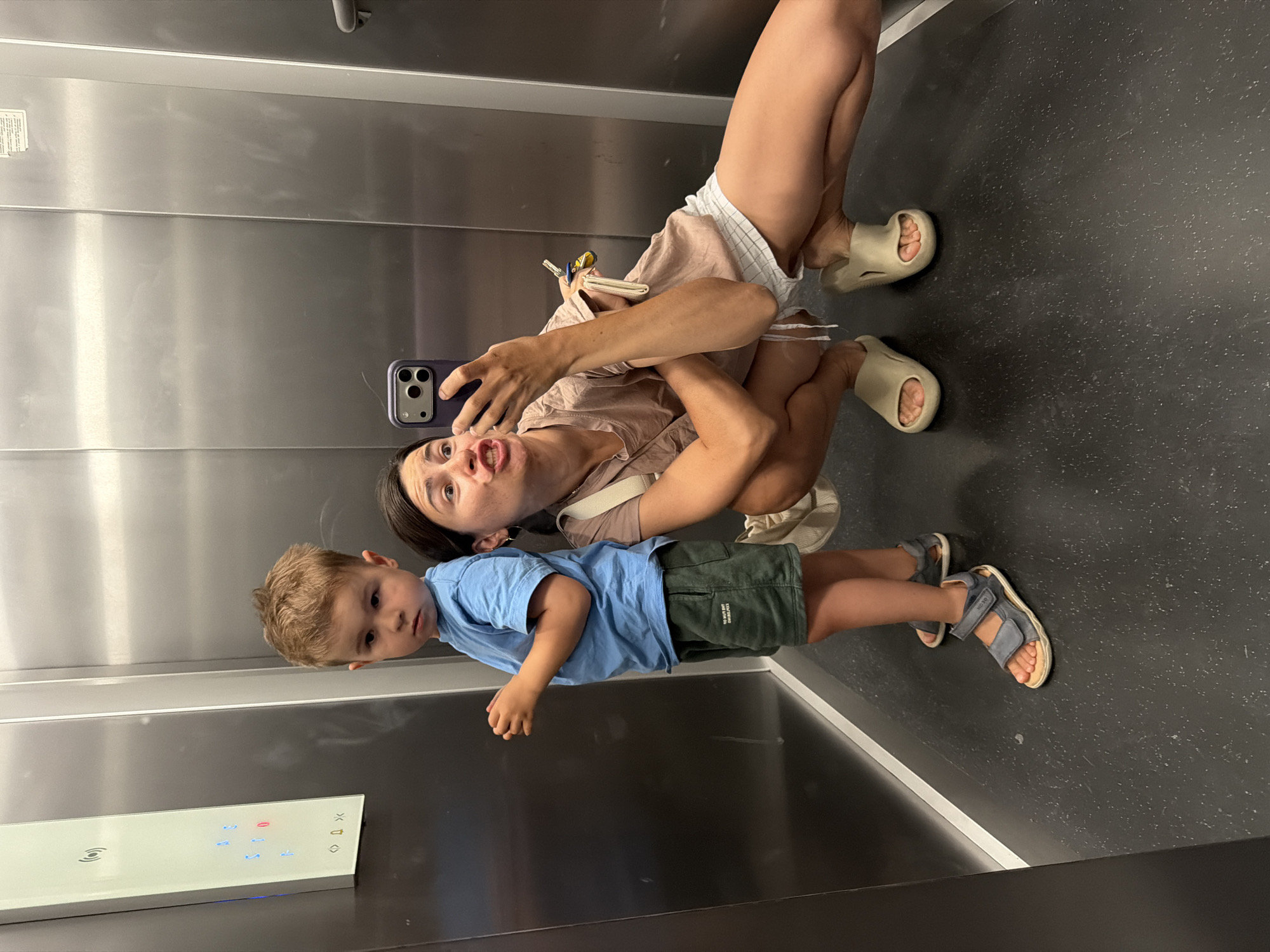 In an elevator, a woman holds a smartphone, presumably taking a selfie with a young boy. The boy, wearing a blue shirt and green shorts, looks towards the camera. The woman wears a light brown top and shorts and beige sliders. The elevator's control panel is visible, suggesting they are inside a building, potentially in Budva, Montenegro, though this cannot be verified from the image. The interior of the elevator is gray and silver.