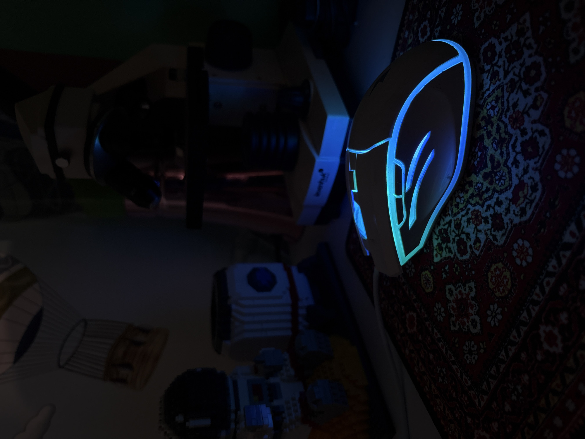 A dimly lit scene shows a gaming mouse with blue and green illuminated edges on a patterned rug. To the left, a microscope and what appears to be Lego bricks are visible. The location is not discernible as Izhevsk, Russia without further information.