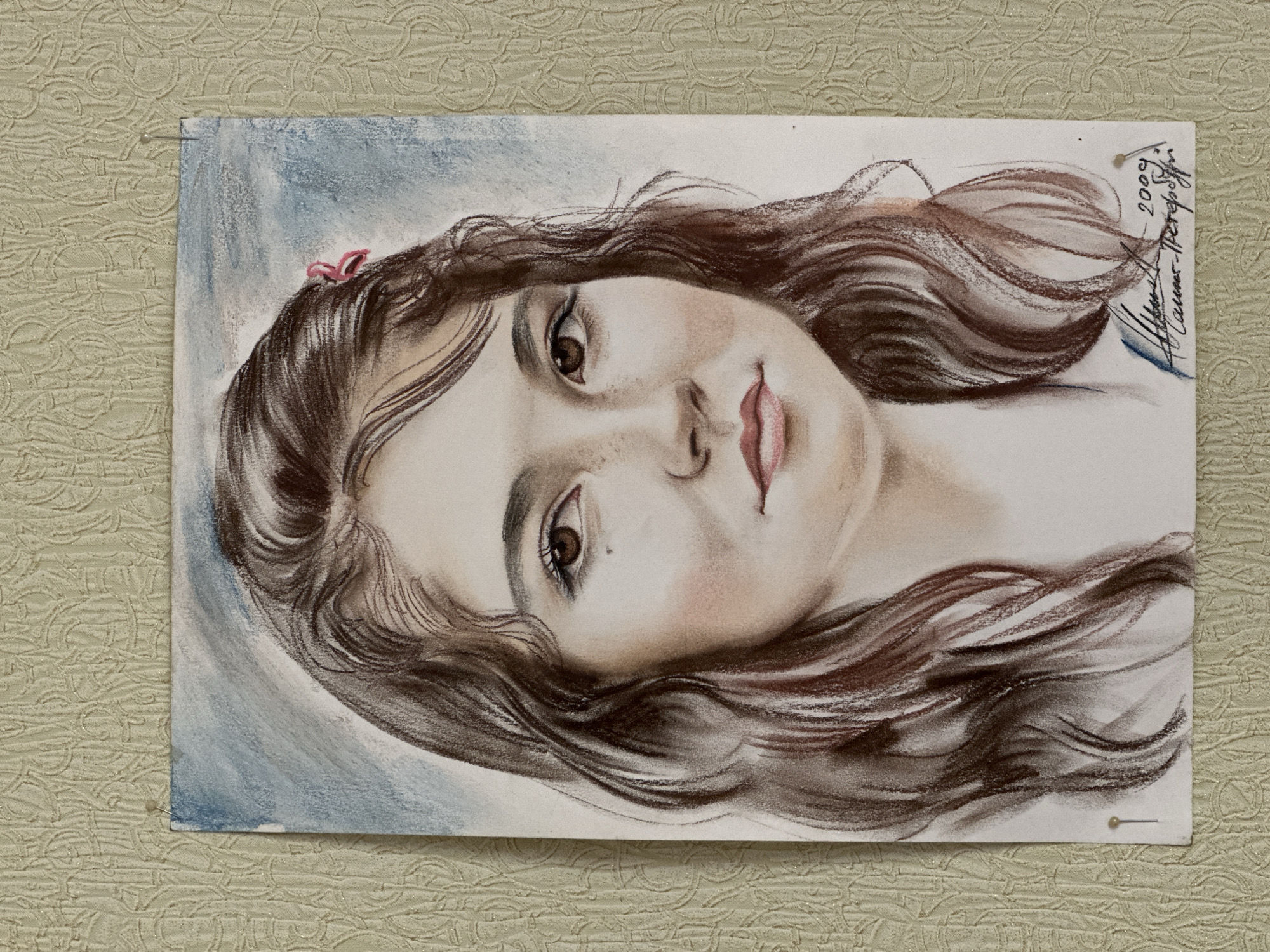 A charcoal and pastel drawing of a young girl's face is pinned to a textured beige wall. The girl has brown wavy hair, light skin, brown eyes, and freckles. A small pink bow is in her hair near the top of the drawing. The background is a soft blue. The artist's signature and the date "2009" are written in the upper right corner of the drawing, along with "Nizhnii Novgorod" written in Russian. Nizhnii Novgorod is a city in Russia.