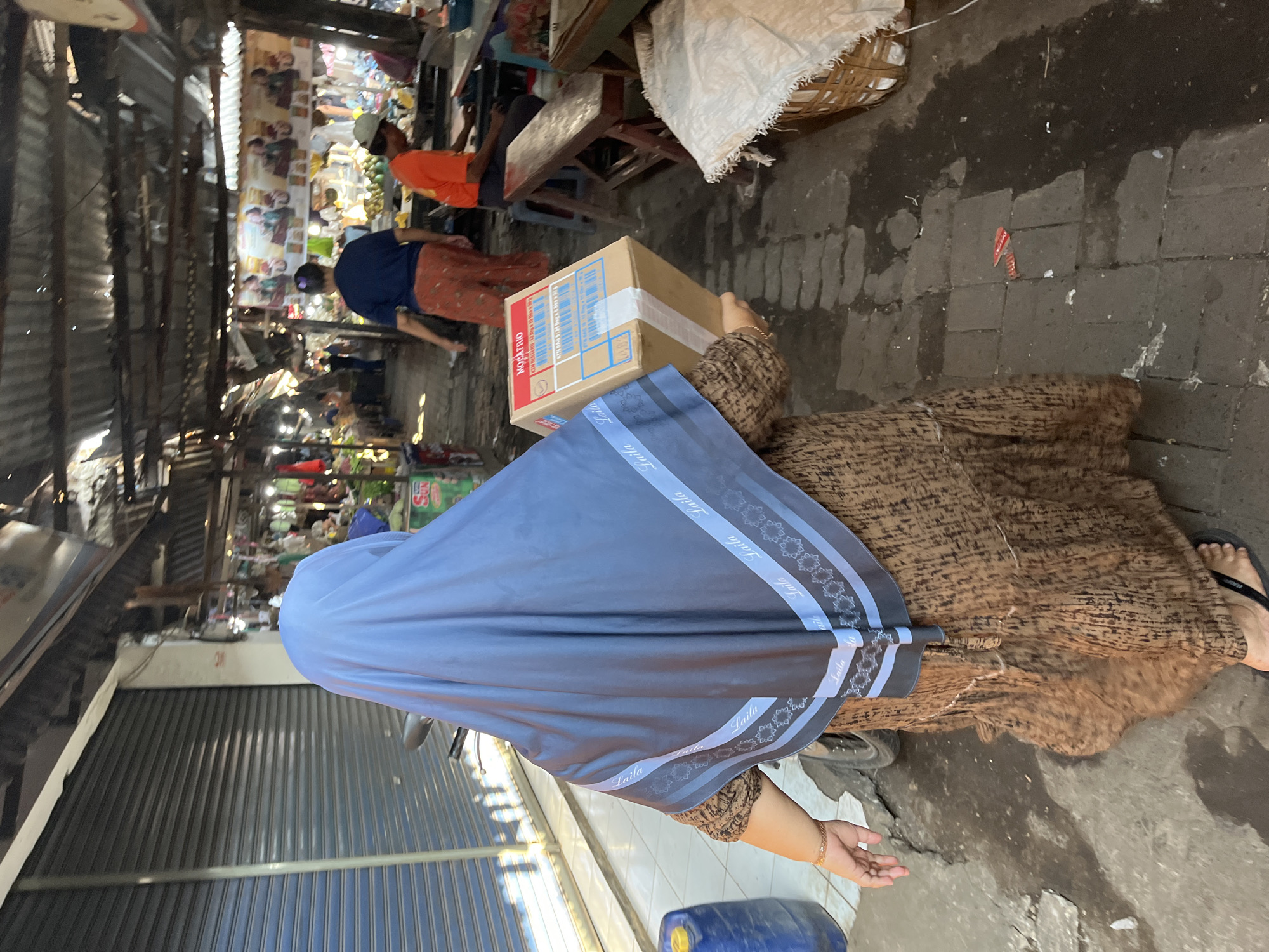 In Larangan, Indonesia, a woman wearing a brown patterned dress and a blue hijab walks down a narrow, covered market aisle carrying a cardboard box. The market has a corrugated metal roof, with stalls on either side selling various goods. The ground is paved with interlocking stones, some of which are wet. In the background, other people can be seen browsing or working in the market. A blue plastic barrel is visible to the left of the woman, and a bicycle is parked next to her. The box she carries is plain brown with some text and labels.