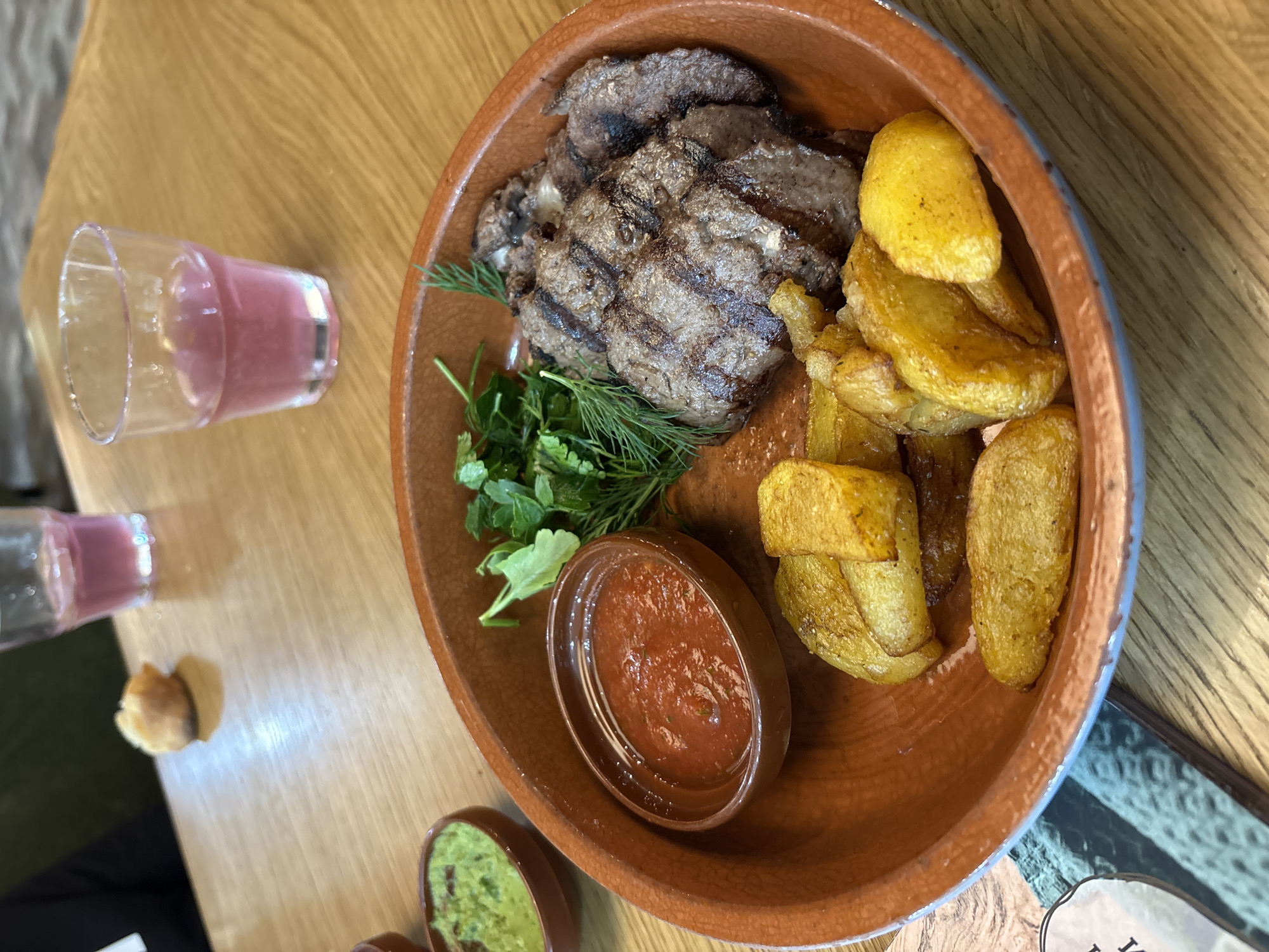 The image shows a brown ceramic bowl filled with a grilled steak cut into slices, fried potato wedges, a small container of red sauce, and fresh herbs, likely dill and parsley. The bowl is placed on a wooden table, likely in a restaurant. A glass of pink liquid is also visible on the table. The presence of additional sauces in small bowls suggests a setting with a focus on dining. The description states it is located in the city of Perm, Russia.