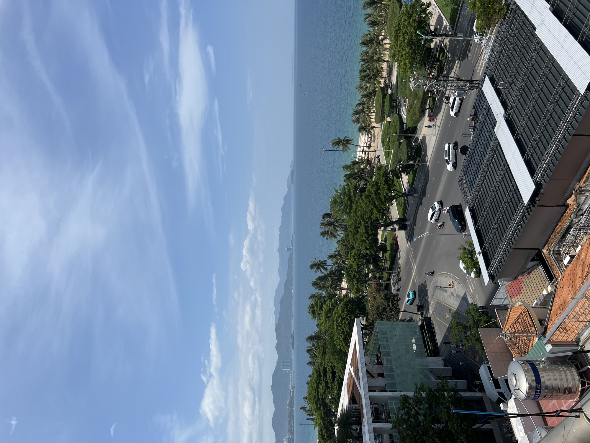The image depicts a coastal urban scene in Nha Trang, Vietnam, from an elevated perspective. The foreground features the rooftops of several buildings, including one with a large silver water tank. Moving towards the midground, a multi-lane road runs parallel to the coastline, with vehicles including cars, a van, and motorcycles in motion. To the right of the road, a well-maintained park area with manicured hedges and palm trees is visible, leading to a sandy beach. Beyond the park and beach, the calm, blue ocean stretches to the horizon. In the distance, a chain of mountains forms a backdrop to the city, with some buildings scattered along the coast. The sky is a clear blue, with scattered white clouds. The overall impression is a vibrant coastal city on a sunny day.