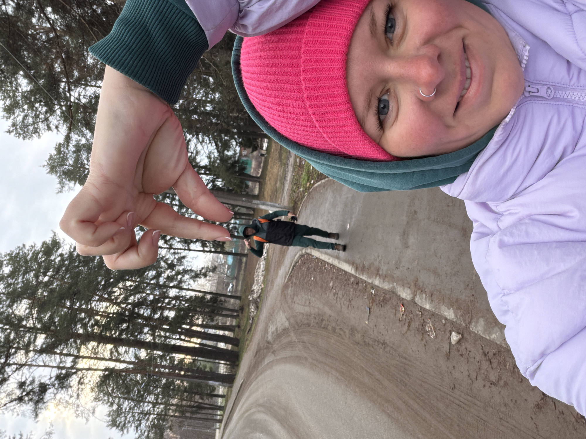 An outdoor selfie captures a person in the foreground utilizing forced perspective to create an optical illusion with another individual in the background. The main subject, positioned on the right and smiling broadly, wears a bright pink knit beanie, a dark green hooded sweatshirt, and a light purple puffer jacket. They have light blue eyes and a silver nose ring. Their hand, held up on the left side of the frame with manicured nails, appears to 'pinch' or hold the smaller person in the distance.
The background figure is a person standing on a paved path, dressed in a dark green tracksuit, possibly with an orange-accented vest or backpack, and dark shoes. Behind them, a line of tall, dark pine trees extends across the frame. The sky is overcast and bright, suggesting daytime with diffused light. The ground is a mix of a paved path and dirt/muddy areas. The setting appears to be a park or an outdoor recreational area in Chaikovsky, Russia. The image conveys a cheerful and playful mood, with no visible text.